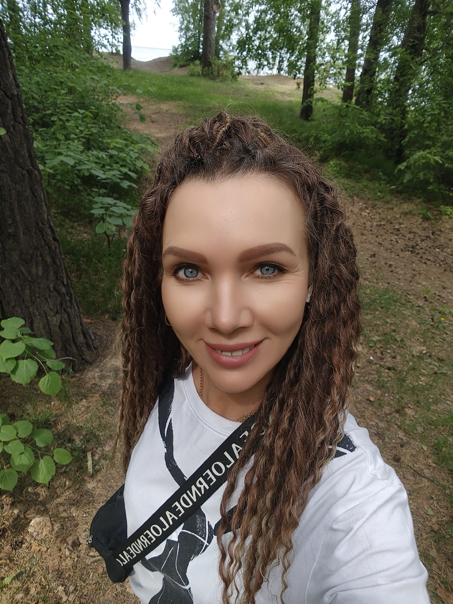 A woman is taking a selfie outdoors in a forested area. She is fair-skinned with blue eyes and light brown, curly hair. She is wearing a white t-shirt with a black graphic design, and a black crossbody bag is slung over her shoulder. The forest is characterized by tall trees, green foliage, and a ground covered in pine needles and grass. In the background, a glimpse of a body of water can be seen through the trees. The location is a wooded area possibly near a lake.