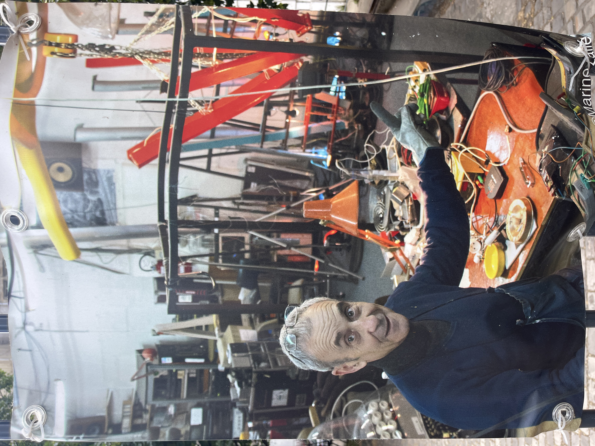 This is a horizontal, slightly tilted photograph of a man in what appears to be a workshop or studio. The man, with gray hair and glasses perched on his head, is looking towards the viewer with a wide smile and open mouth. He is wearing a dark blue top. His right arm is raised, and he is wearing a light gray glove, suggesting he might be in the process of working on something or demonstrating a tool.
The background is cluttered with various objects, including metal structures, chains, tools, wires, and electronic components. Bright red and yellow elements are interspersed among the darker equipment, adding visual interest. The overall impression of the setting is one of a busy, functional space, possibly for art, construction, or repair. The lighting appears to be artificial, casting shadows and illuminating the scene. There are some white grommets visible along the bottom and left edges of the image, indicating it might be a banner or part of a larger display.
A small amount of text is visible vertically on the right side of the image, appearing to read "@Marine_Lane". The time of day and weather are not discernible from the image. The man's expression conveys enthusiasm and engagement.