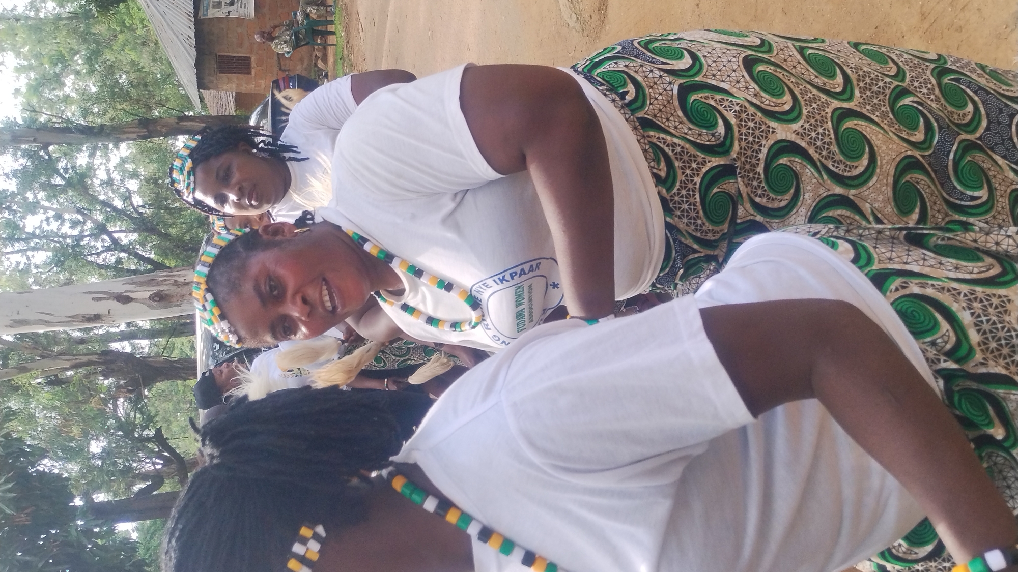 A medium shot captures three Nigerian women in traditional attire during what appears to be a celebration or cultural event in Jos, Nigeria. The woman in the foreground, facing right, wears a white t-shirt, a beaded headband adorned with yellow, green, and white beads, and a matching beaded necklace. She also wears a patterned skirt with green and black swirls on a beige background. Her mouth is open as if speaking or laughing. Behind her, a woman also wearing a white t-shirt and a beaded headband with similar colors looks to her right. A third woman, slightly further back and to the right, wears a white t-shirt and a beaded headband. The background shows trees, a rustic building with a corrugated iron roof, and a dirt road. Another person sits on a chair in the background. The scene is outdoors and appears to be a public gathering.