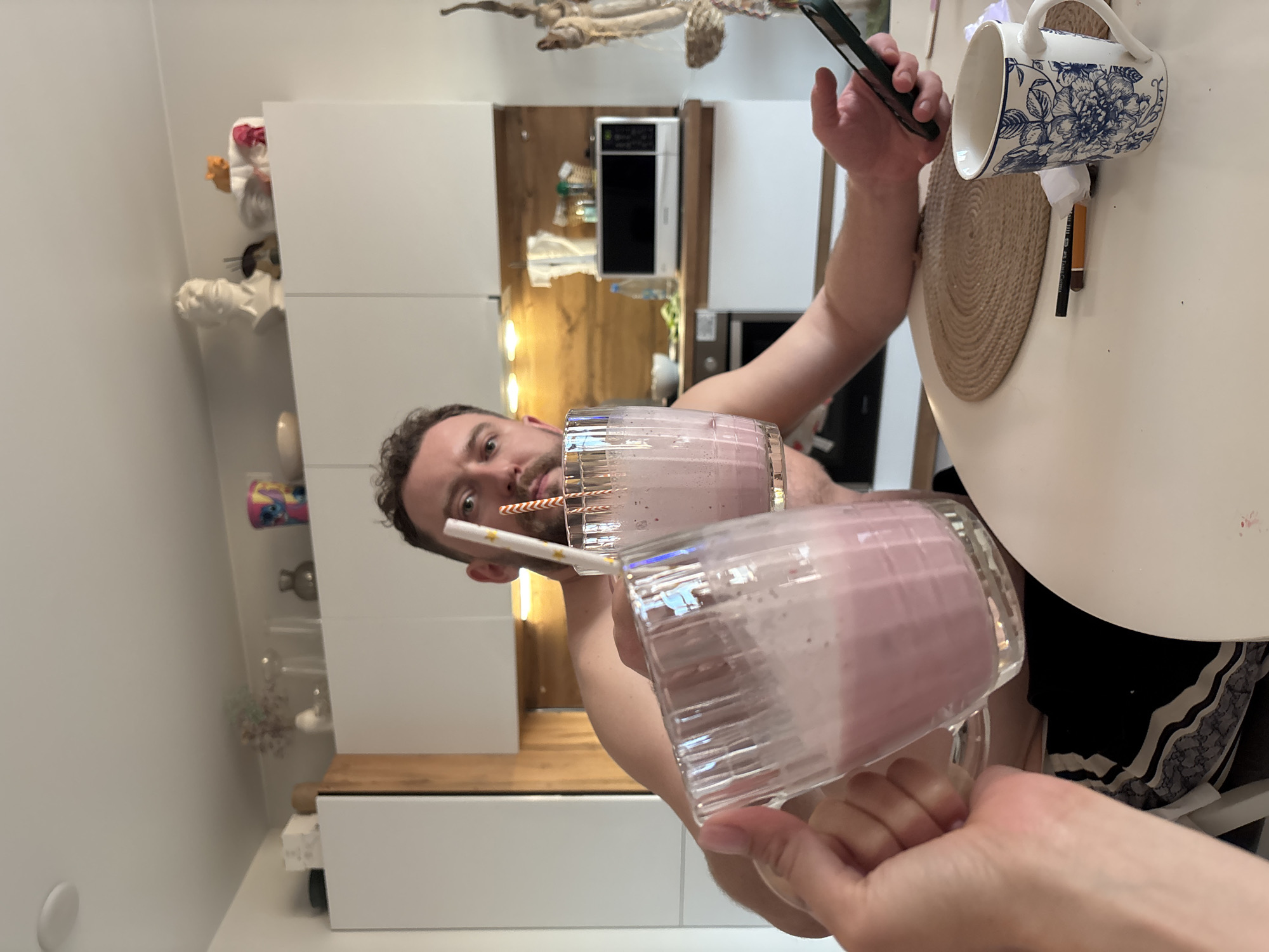 A bright indoor photograph, taken in a modern kitchen or dining area in Krasnoyarsk, Russia, captures two individuals making a toast with pink, frothy milkshakes or smoothies.
In the mid-ground, a man with short, dark curly hair and a beard looks directly at the camera with a neutral expression, holding a clear glass mug with a red and white striped straw in his left hand. His right hand holds a smartphone with a dark case. In the immediate foreground, the hands of another person are visible, holding a second identical glass mug containing the pink beverage, complete with a white straw adorned with gold stars. Their hands are positioned as if clinking glasses for a toast.
The setting features a white table, part of which is visible, with a round braided placemat. On the table, there is a white mug with a blue floral pattern and a dark marker or pen. The background reveals a clean, contemporary kitchen space with white cabinets, wooden wall panels or countertops, and a silver microwave oven. Various decorative items are arranged on shelves or cabinet tops, including a white ceramic figurine, a small orange object, and a colorful mug potentially featuring the character Stitch.
The lighting is bright and even, suggesting an indoor daytime setting. The overall mood is casual and friendly. No visible text is discernible in the image.