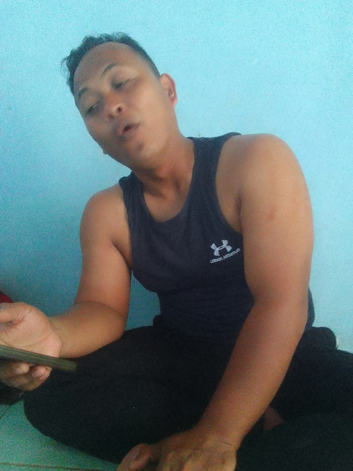 A young man of East Asian descent with dark hair and brown eyes is shown from the chest up, sitting on the floor with a light blue wall behind him. He is wearing a dark blue Under Armour tank top and dark pants. His left arm is bent, and he is holding a flat, rectangular object, possibly a phone or a book, in his left hand. His head is tilted back slightly, and his mouth is open as if he is speaking or singing. The lighting is dim, casting a shadow on the right side of his body. The scene appears to be an indoor setting, likely a room in a house or apartment in Suren, Indonesia.