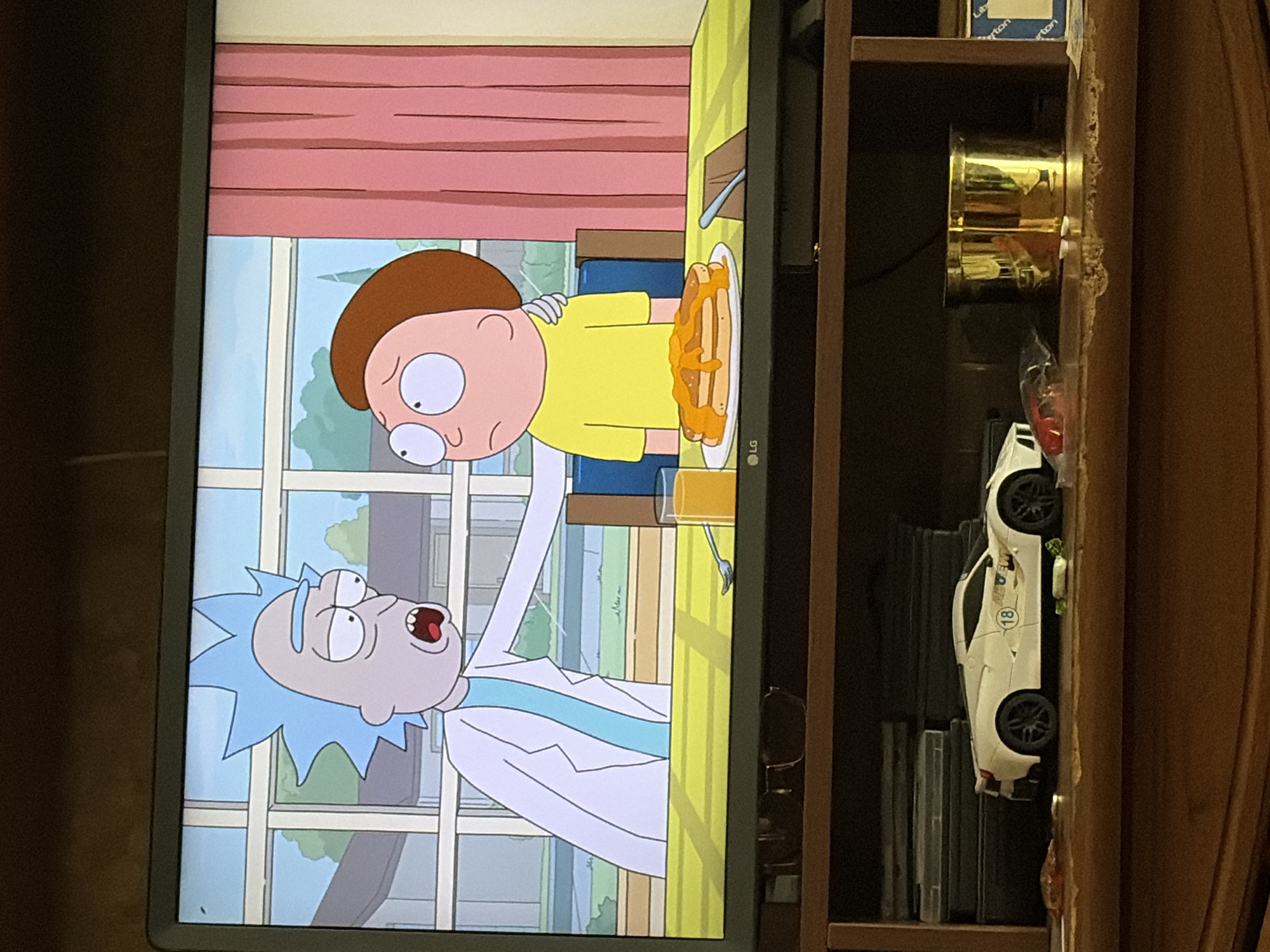 The image depicts a television screen displaying an animated scene from the TV show "Rick and Morty." Rick Sanchez, with his signature blue spiky hair, is seen with his hand on Morty Smith's shoulder. Morty, wearing a yellow shirt, looks slightly distressed. On the table in front of them are pancakes drenched in syrup and a glass of orange juice. The background shows a domestic setting with windows and pink curtains. Below the television, on a wooden shelf, are various items including a white toy car, stacks of DVDs, a golden container, and other miscellaneous objects. The overall scene is captured indoors, likely in a living room or entertainment area.