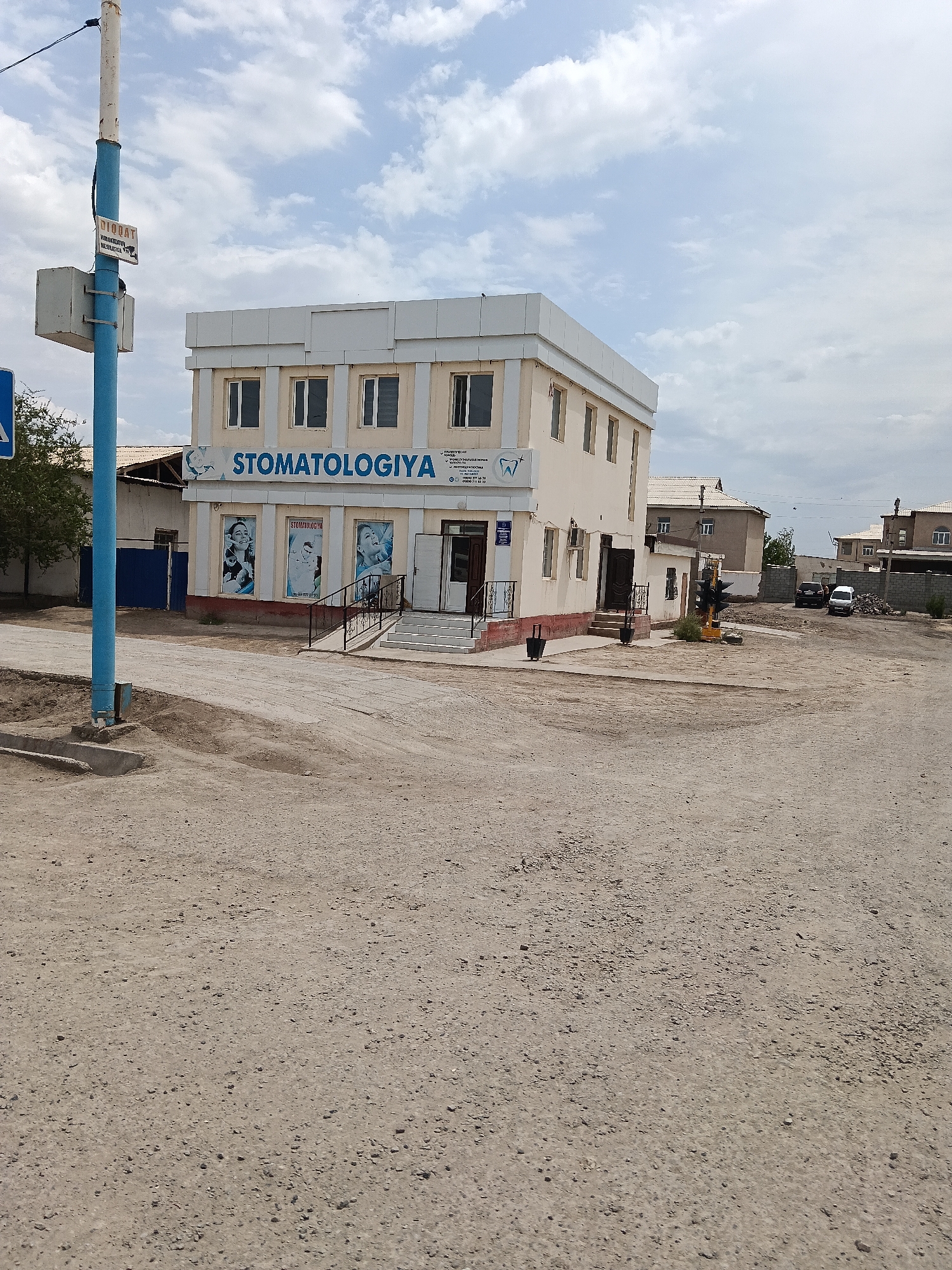 The image shows a two-story building with the sign "STOMATOLOGIYA" on the upper level, indicating it is a dental clinic. The building is light beige with white trim and multiple windows. In front of the building, there is a paved entrance with stairs and a railing. Three posters are visible on the lower level, two of which appear to show images of smiling people with healthy teeth. A blue lamppost with a white sign is located to the left of the building. The scene appears to be in a semi-urban or rural setting, with unpaved ground and a few other buildings in the background. The sky is partly cloudy. The location is identified as the city of Kungrad, Uzbekistan. There are no people visible in the image.