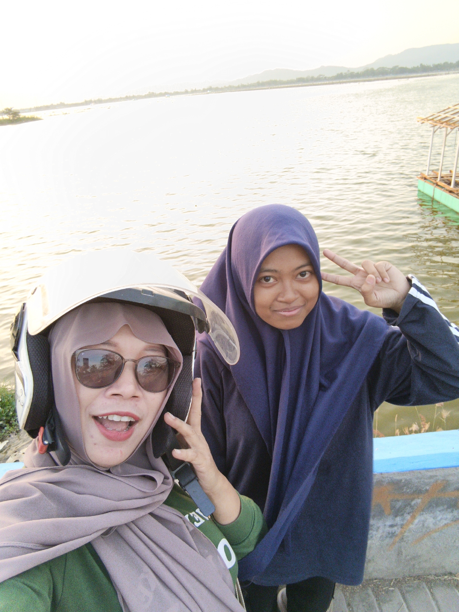 Two young women are taking a selfie by a body of water in Krakitan, Indonesia. The woman on the left is wearing a white helmet with a clear visor, sunglasses, and a light purple hijab. She is also wearing a green long-sleeved shirt. Her mouth is open and she is smiling. The woman on the right is wearing a dark blue hijab and a dark blue long-sleeved shirt with white stripes on the sleeves. She is holding up two fingers in a peace sign. The background shows a body of water with a small raft or boat with a thatched roof visible in the upper right corner. There are hills and mountains in the distance. The lighting suggests it is late afternoon or early evening.