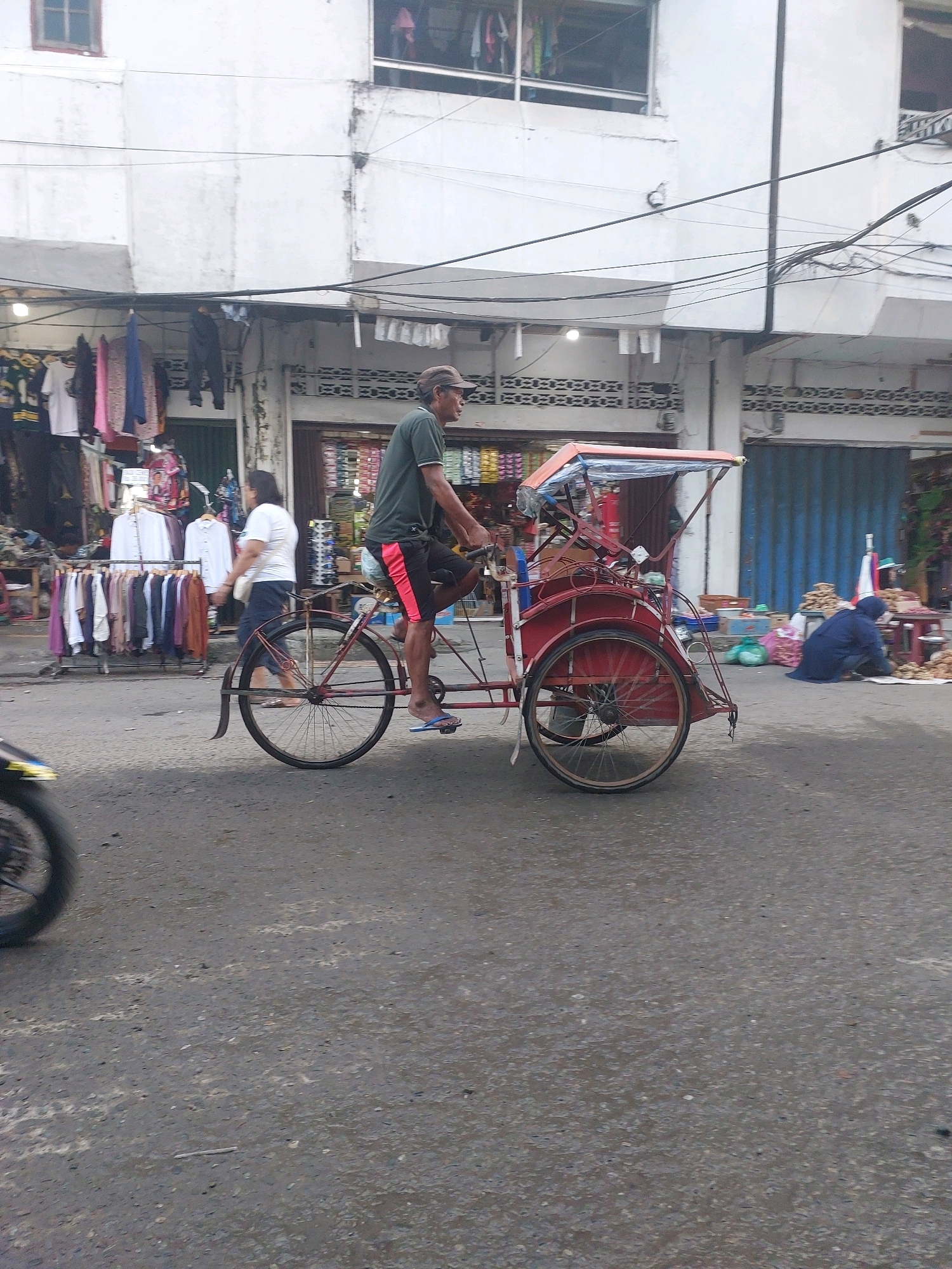 The image shows a street scene in Ambon, Indonesia, likely on a commercial street given the presence of storefronts and vendors. A man is riding a pedicab, a three-wheeled vehicle powered by pedaling, with a covered passenger area at the rear. He is wearing a dark green t-shirt and red and black shorts. Another person, a woman, is walking away from the pedicab towards a row of clothing racks displayed outside a shop. The buildings in the background appear to be multi-story structures with awnings and open storefronts. Electrical wires are strung across the street overhead. In the background, a person in a blue garment is crouching near a market stall with produce and goods. The street is paved and appears wet in some areas, suggesting recent rain or a cleaning effort. Other vehicles, including a motorcycle wheel in the foreground left, are partially visible. The overall impression is one of a busy, everyday scene in a tropical urban environment.