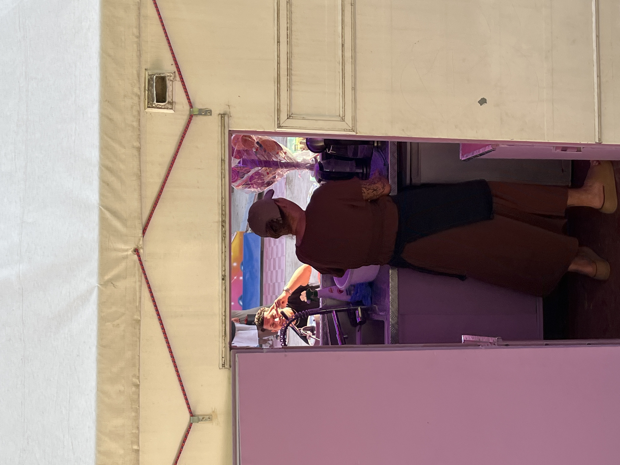 A medium shot shows the interior of a food stall, possibly at an outdoor event, with two people visible. A woman wearing a brown t-shirt, an apron, and tan slides stands facing a counter and appears to be working. She has a tattoo on her left arm. A man in a black t-shirt and a baseball cap is seen through an opening in the stall, holding up two fingers in a peace sign. He is smiling and looking at the camera. The stall is decorated with large, colorful lollipops in the background. The stall itself appears to be made of white and beige material, with red bungee cords securing a section of the awning. The floor inside the stall is dark. The image does not provide any specific visual cues to indicate its location in Paris, France.