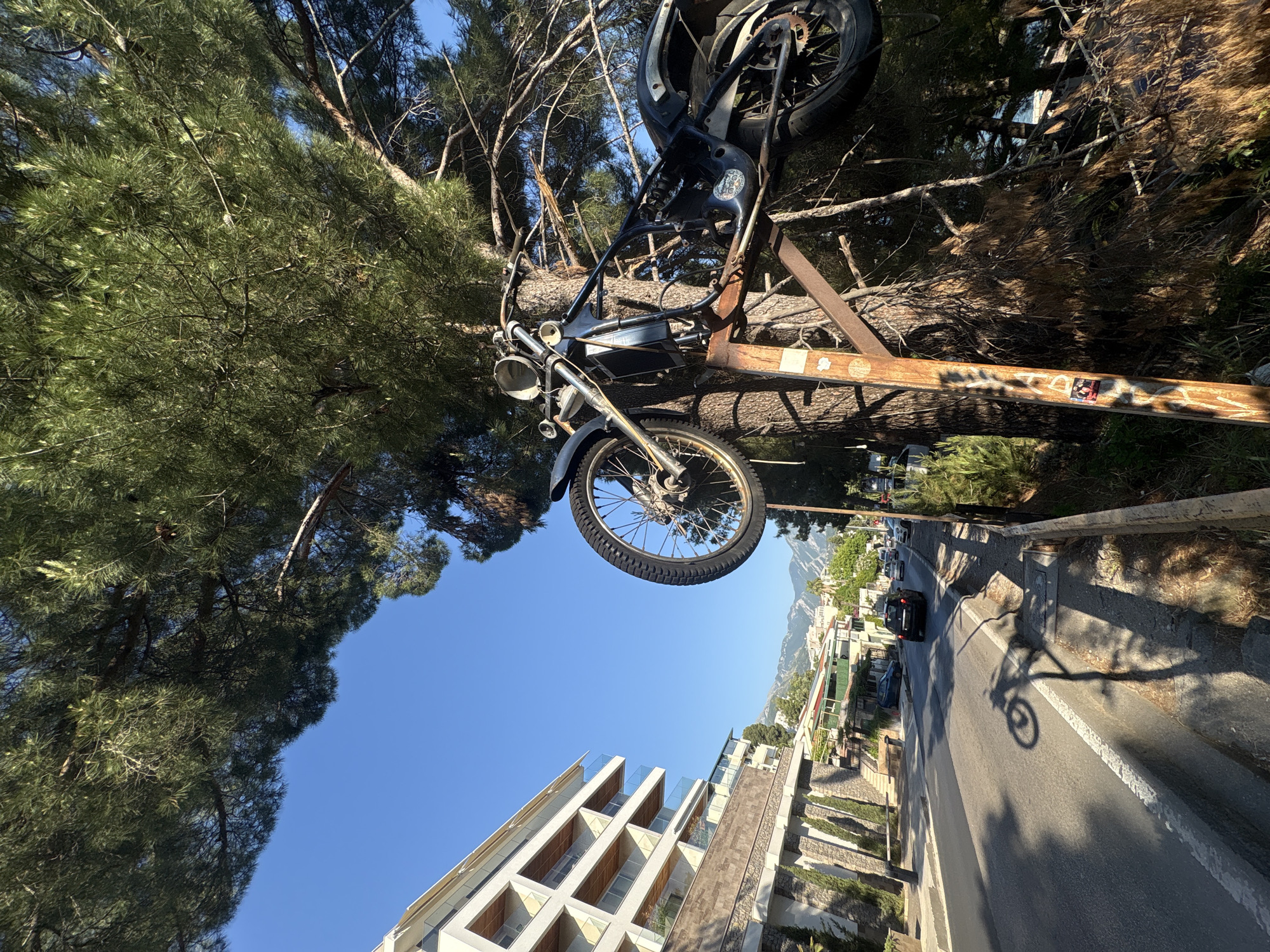The image shows a motorcycle mounted on a rusty metal frame amongst trees, overlooking a street in Bar, Montenegro. A modern multi-story building is visible in the lower left of the frame. A road with vehicles is on the right, with mountains in the distance. A long shadow of the motorcycle is on the street. The sky is clear and blue.