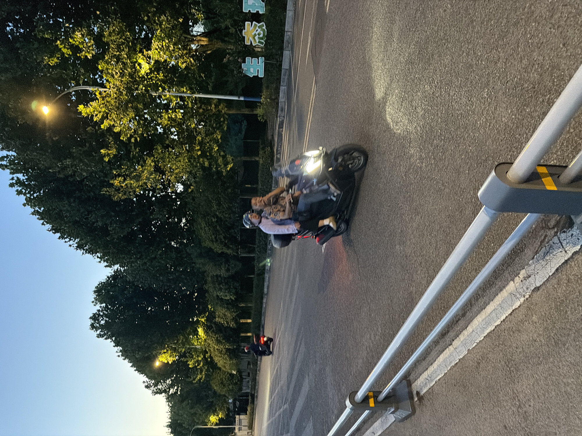 A street scene at dusk in Jiangjin, China, features two people riding a scooter on a paved road. The scooter is black, with the driver wearing a patterned shirt and a helmet. A passenger, also wearing a helmet, sits behind the driver. A pedestrian crossing is visible in the background, along with another scooter further down the road. Streetlights illuminate the scene, casting a warm glow on the trees lining the street. A sign with Chinese characters is visible on the right side of the image. A metal guardrail runs along the left side of the road. The sky is clear and transitioning from blue to a lighter hue, indicating twilight.
