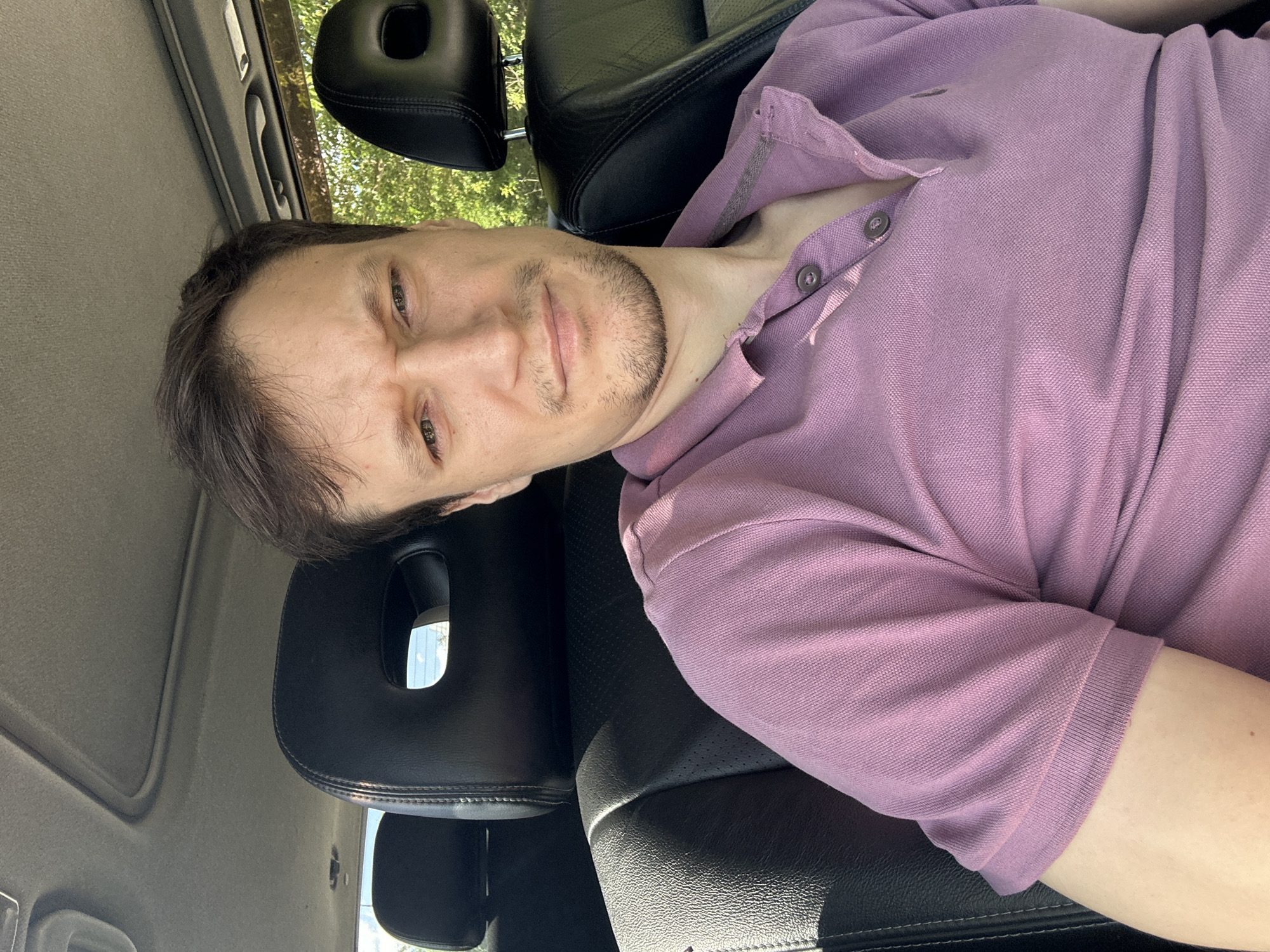 A man with brown hair and a light beard is sitting in the driver's seat of a car, looking directly at the camera. He is wearing a light purple polo shirt with two buttons at the neck. The interior of the car is visible, including black leather seats with cutouts in the headrests and a sunroof. Sunlight filters into the car from outside, illuminating the scene. The background appears to be a natural setting with trees and foliage. The image was taken in Makiv, Ukraine.