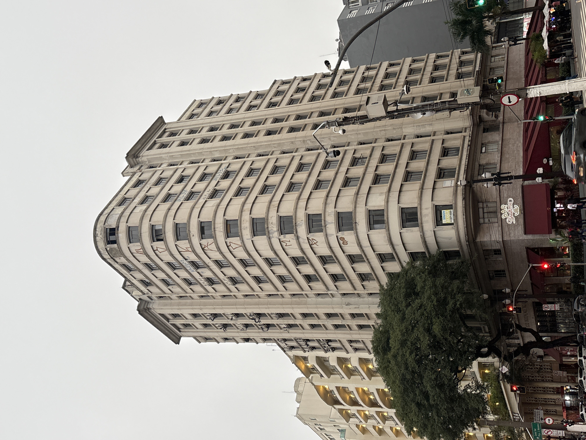 This is an eye-level, outdoor, daytime photograph of the Edifício Planalto, a historic building located in the city of São Paulo, Brazil, specifically at the intersection of Rua Álvares Penteado and Rua XV de Novembro in the historic downtown area. The building, an example of Art Deco architecture, features a distinctive curved facade with multiple stories and numerous windows. Its exterior is a light beige color, and some of the windows appear to be damaged or boarded up, with graffiti visible on the facade of the upper floors.
In the foreground, to the left, a large tree with dark green foliage partially obscures the lower floors of the building. Red awnings extend from businesses on the ground floor, including one with a sign that reads "BAR BRAHMA". Traffic lights are visible, with at least one showing red and another green. Street lamps and electrical poles with security cameras are also present.
To the left of the Edifício Planalto, another building with a cream-colored facade and arched balconies, illuminated from within, is visible. Further in the background, a darker, more modern building rises, contrasting with the older architecture. Several cars are on the street, with a dark gray SUV in motion in the lower right portion of the frame. Pedestrians are visible on the sidewalks and crossing the street, dressed in casual clothing. The sky is overcast, contributing to a muted lighting condition.
