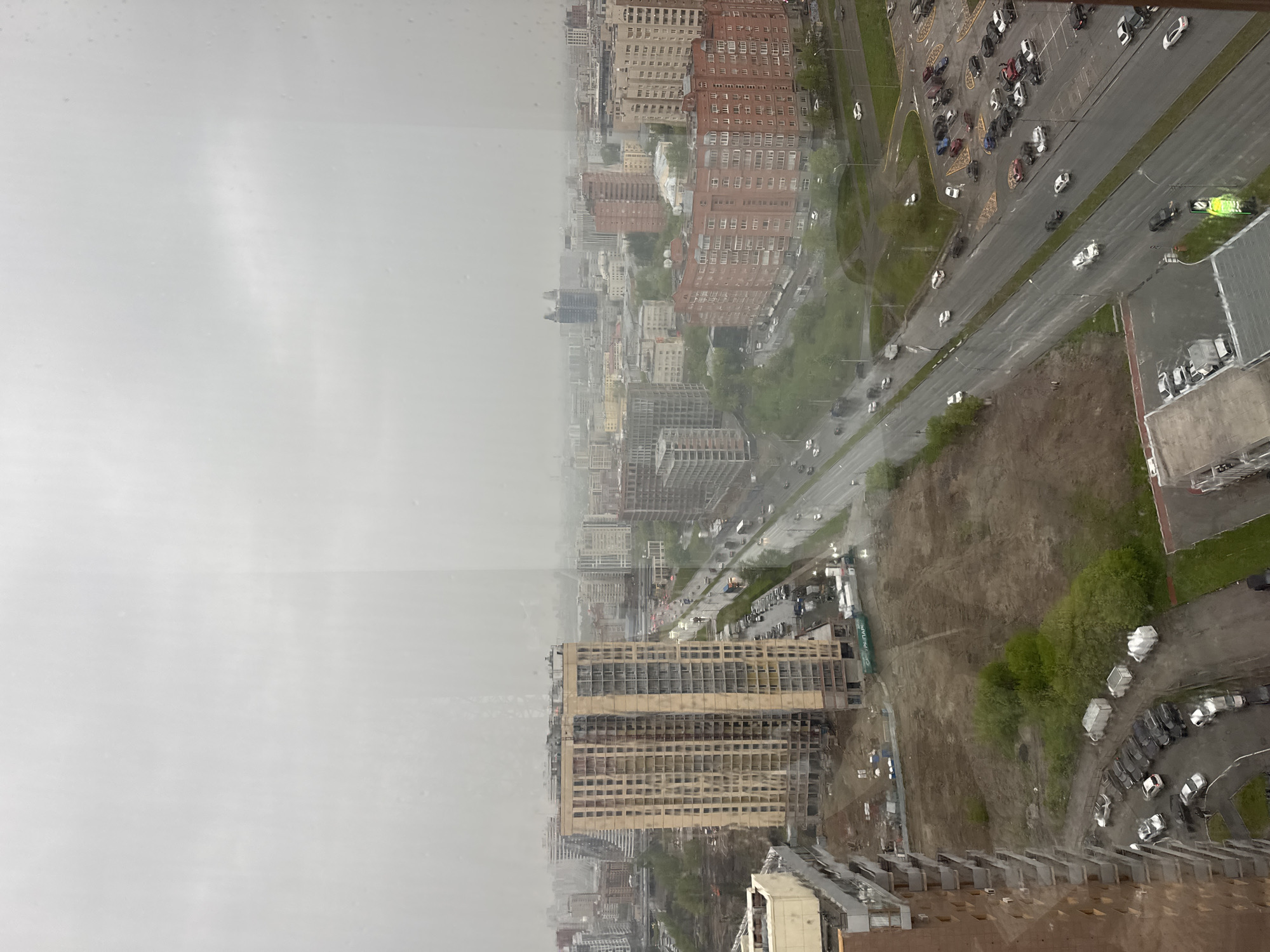 An aerial view of Novosibirsk, Russia, under heavy rain and overcast skies. Predominantly grey, the urban landscape features a mix of buildings ranging from older red-brick structures to modern high-rises in varying states of construction. Dense building clusters fade into the distance due to the rain and atmospheric haze. A major road bisects the scene, showing visible car traffic. Adjacent to the road is a bare, muddy area dotted with some green vegetation and parked cars. A partially completed high-rise building is in the foreground. The view is captured through a rain-streaked window.