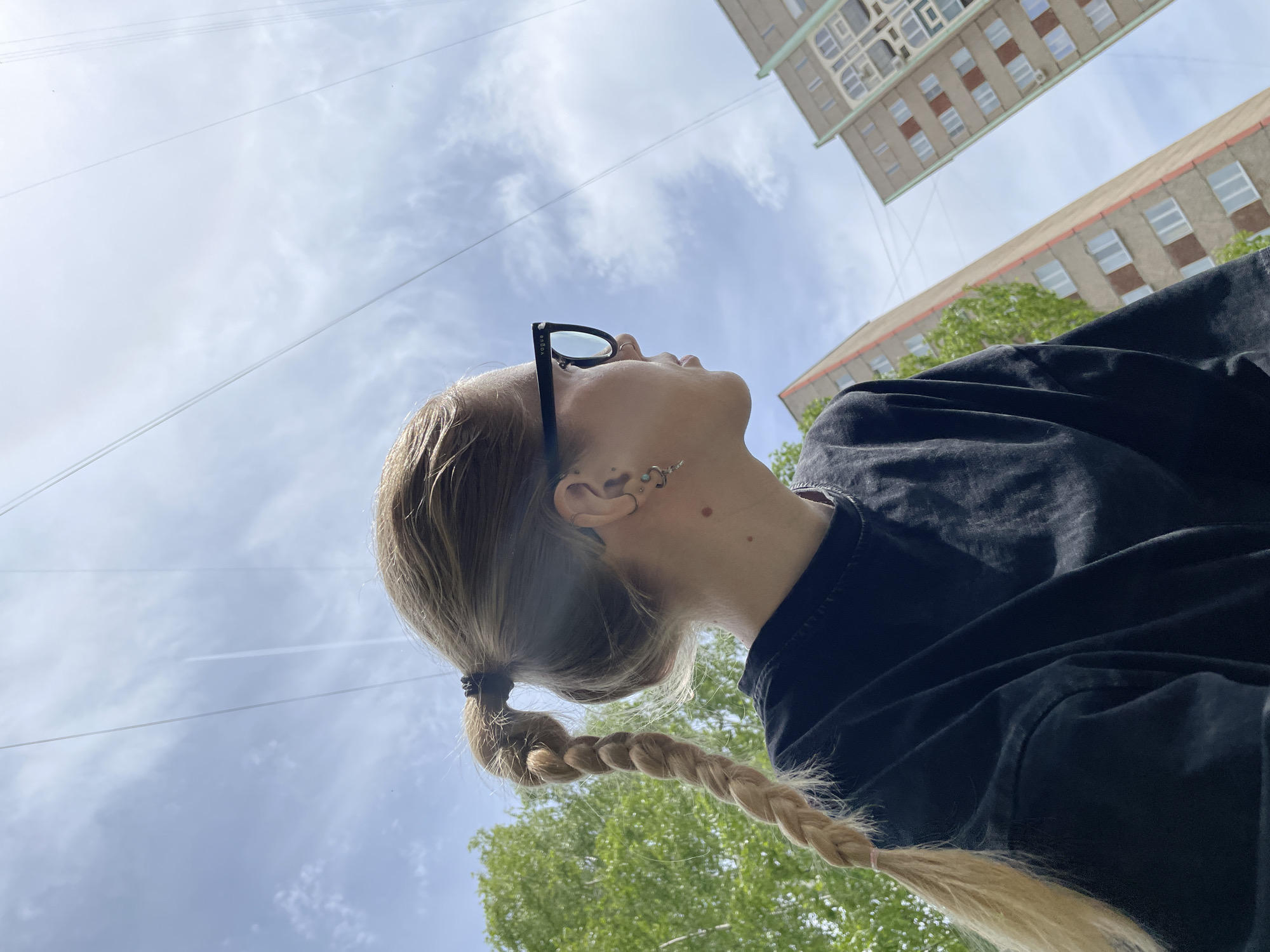 The photo is taken from a low angle, looking up at a woman with blonde hair in a braid, wearing glasses and a dark shirt. She is looking upwards towards a sky with scattered clouds. Behind her are two buildings, one taller than the other, with multiple windows, and some green trees. The buildings are located in the city of Izhevsk, Russia. There are also some power lines visible against the sky.