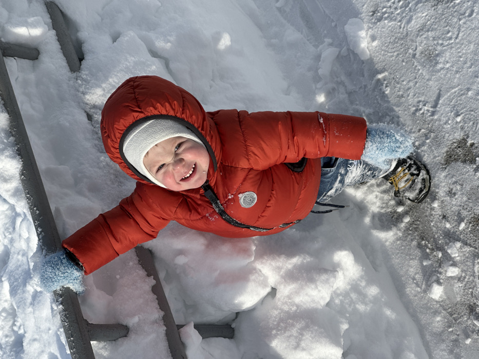 A young child, appearing to be a toddler, is observed in a snowy outdoor environment in Perm, Russia. The subject is dressed in a red hooded puffer jacket with a circular, silvery emblem on the left chest, a white fleece beanie, blue denim pants, light blue mittens, and black-and-yellow winter boots. The child's left hand, covered by a mitten, grips a horizontal metal rail that is partially obscured by snow. A substantial snowdrift extends along the rail to the child's left. The ground immediately beneath the child's feet consists of a partially cleared surface with scattered snow and ice. The child is looking upwards toward the camera, displaying a wide smile with crinkled eyes. The scene is brightly illuminated by direct sunlight, indicating a clear winter day.