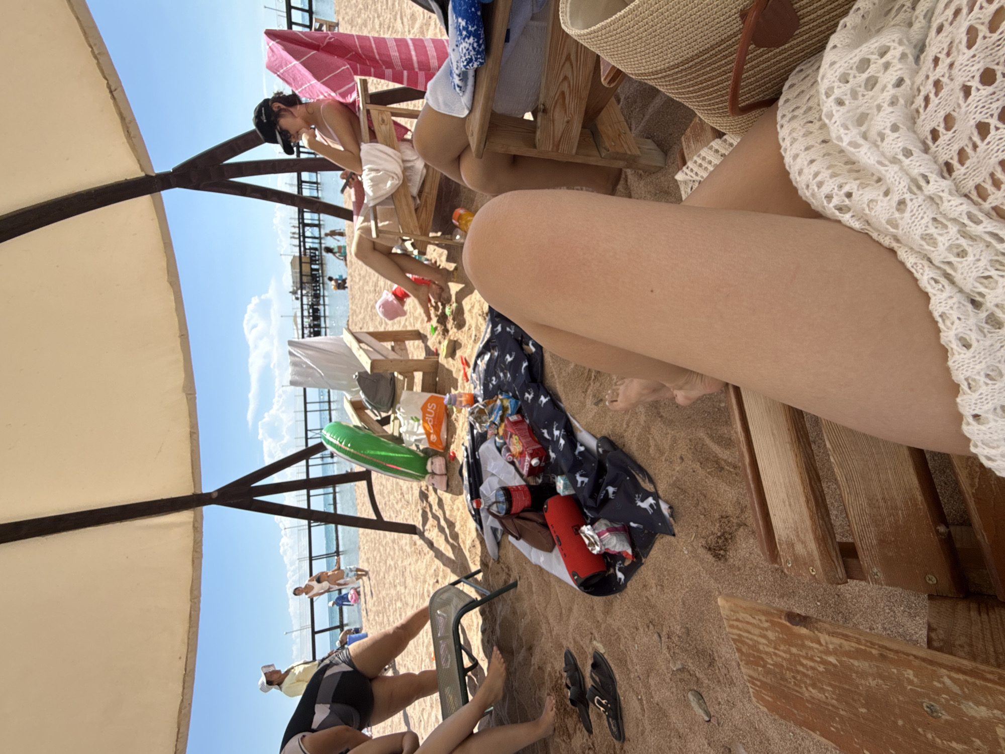 A group of people are relaxing on a sandy beach under a large, light-colored canopy. The scene appears to be in Sary-Oy, Kyrgyzstan, given the context provided. In the foreground, a person's legs are visible, draped with a white, crocheted cover-up. To the left, a low lounge chair is occupied by at least one individual, with sandals resting on the sand nearby. A blanket spread on the sand is adorned with a pattern of deer and covered with various items including a red speaker, a bottle of Coca-Cola, snacks in bags, and a red box. Further back, a woman sits in a wooden lounge chair, wearing sunglasses and a cap, and holding a phone. Another person is visible in a similar chair. In the background, children are playing on the sand, and a pier extends into the blue water under a bright sky with scattered clouds. There are inflatable toys, a pink towel, and a straw bag present among the beach accessories. The overall atmosphere suggests a leisure outing at the beach.