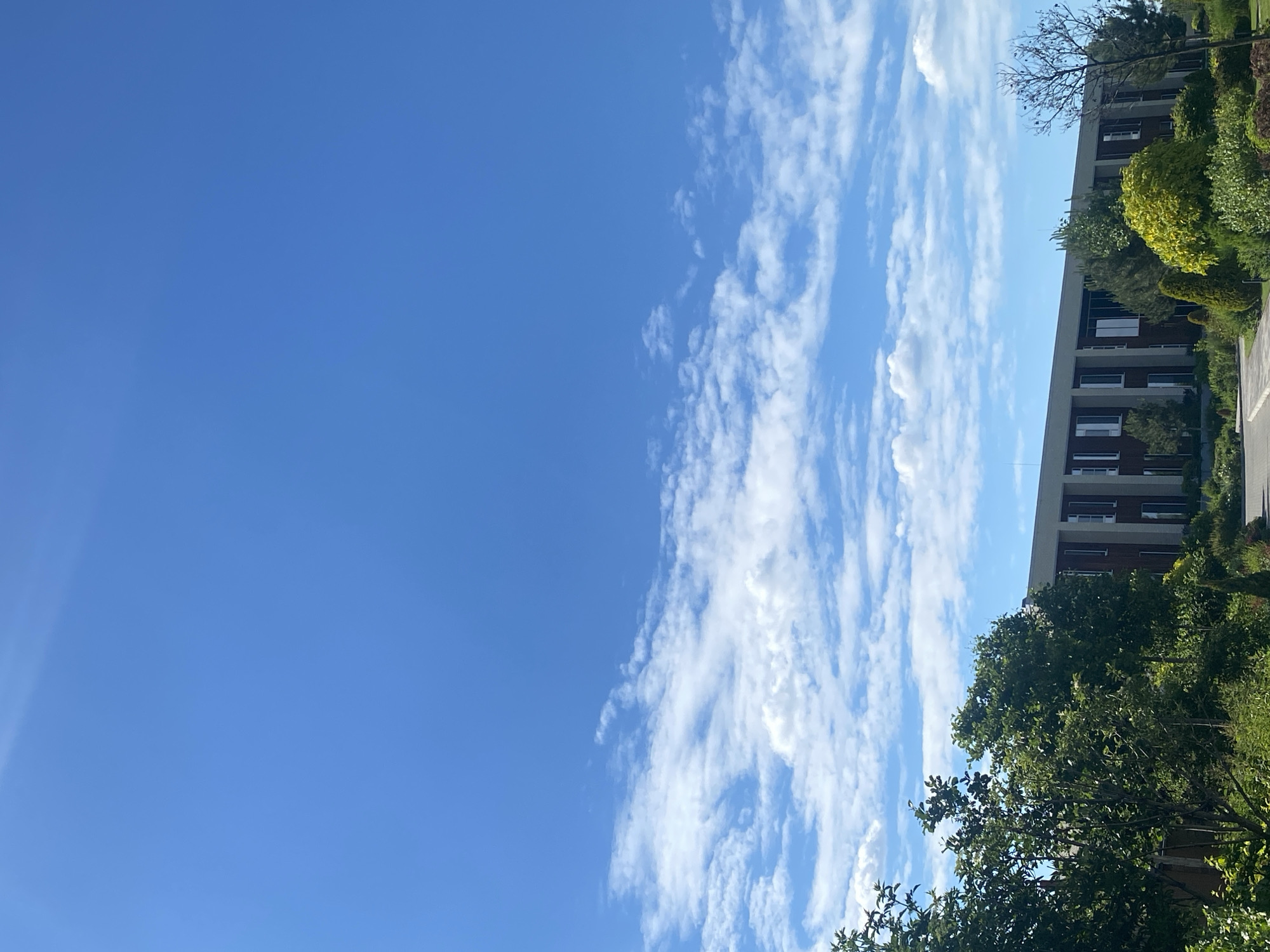 The image depicts a modern, multi-story building with a brick facade and a light-colored frame structure, observed from a distance. The building has a long, rectangular shape and is flanked by mature trees and lush green landscaping. The sky above is a vibrant, clear blue with scattered white, wispy clouds. The scene appears to be set in a suburban or semi-rural area, possibly a business park or an educational institution, given the architecture and well-maintained grounds. The overall impression is one of a bright, sunny day with good visibility. The location is identified as Rozhny, Ukraine. There are no people or animals visible in the image, and no actions or interactions are taking place. The focus is on the architectural structure, the natural surroundings, and the expansive sky.