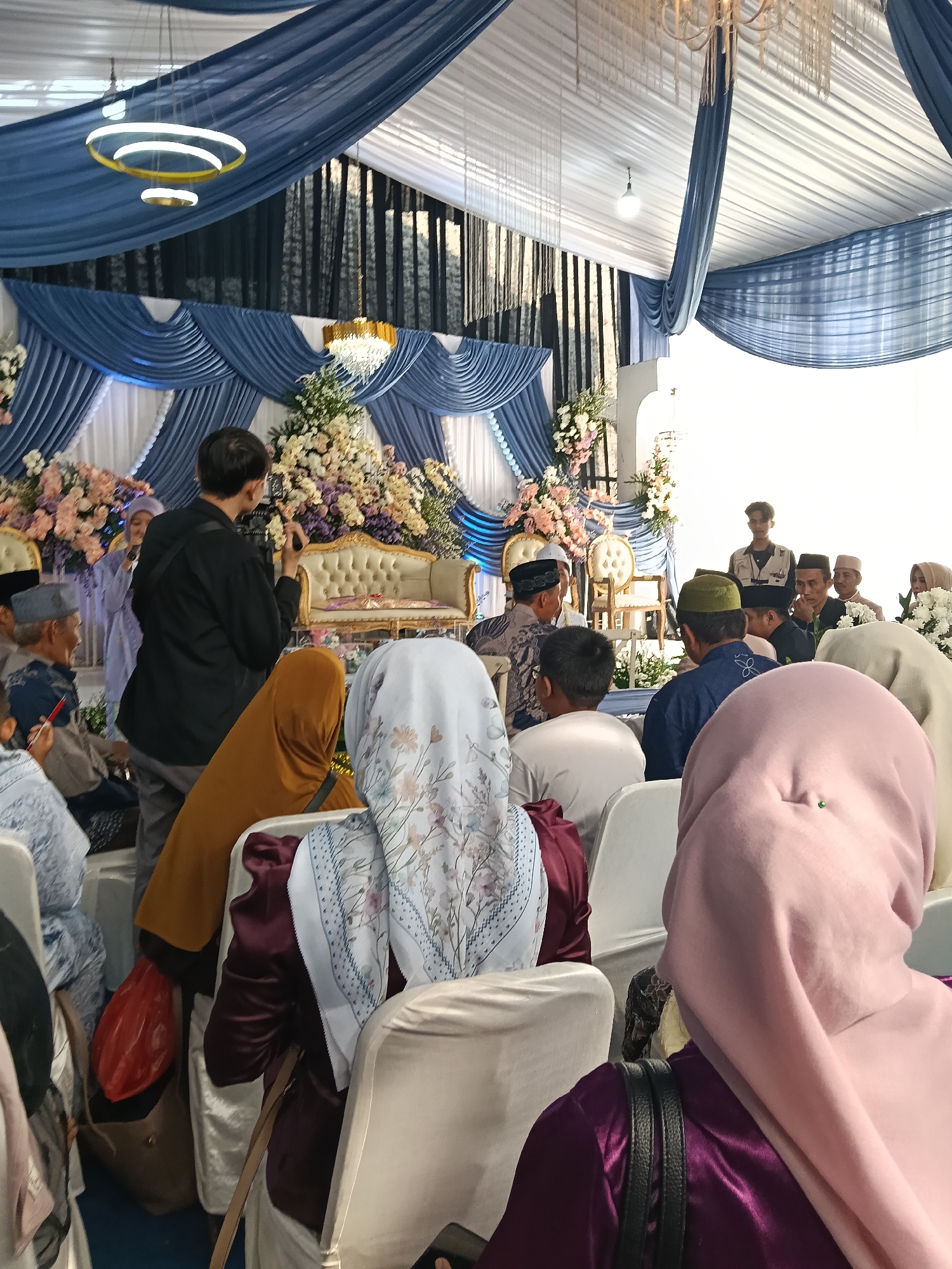 The image depicts an indoor event, likely a wedding ceremony, in Tegal Angus, Indonesia. A man in a black shirt is filming with a camera in the foreground. Several people are seated on chairs, with many wearing religious head coverings. A decorated sofa and floral arrangements suggest a ceremonial setting. Draping fabric in blue and white adorns the ceiling, and modern chandeliers hang overhead. The overall atmosphere appears to be formal and celebratory.