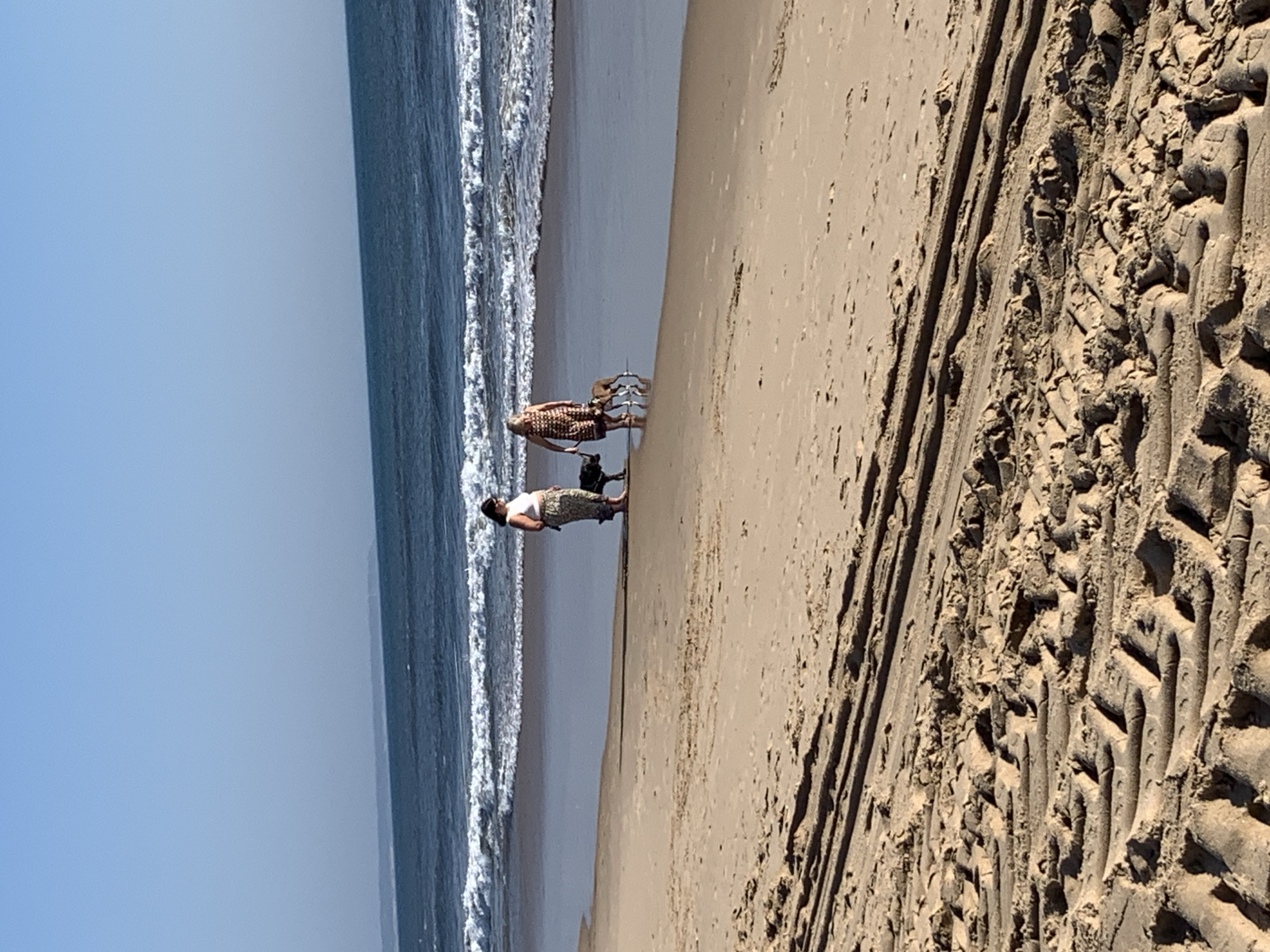 Two individuals and three dogs are walking on a sandy beach. The person on the left wears a white top and patterned pants, and is accompanied by a dark-colored dog. The person on the right wears a patterned dress and walks with two dogs, one dark-colored and one lighter brown. The ocean is visible in the background with waves breaking on the shore. The foreground shows tire tracks in the sand. This scene is consistent with the beaches of Costa da Caparica, Portugal, known for their extensive sandy shores and recreational activities.