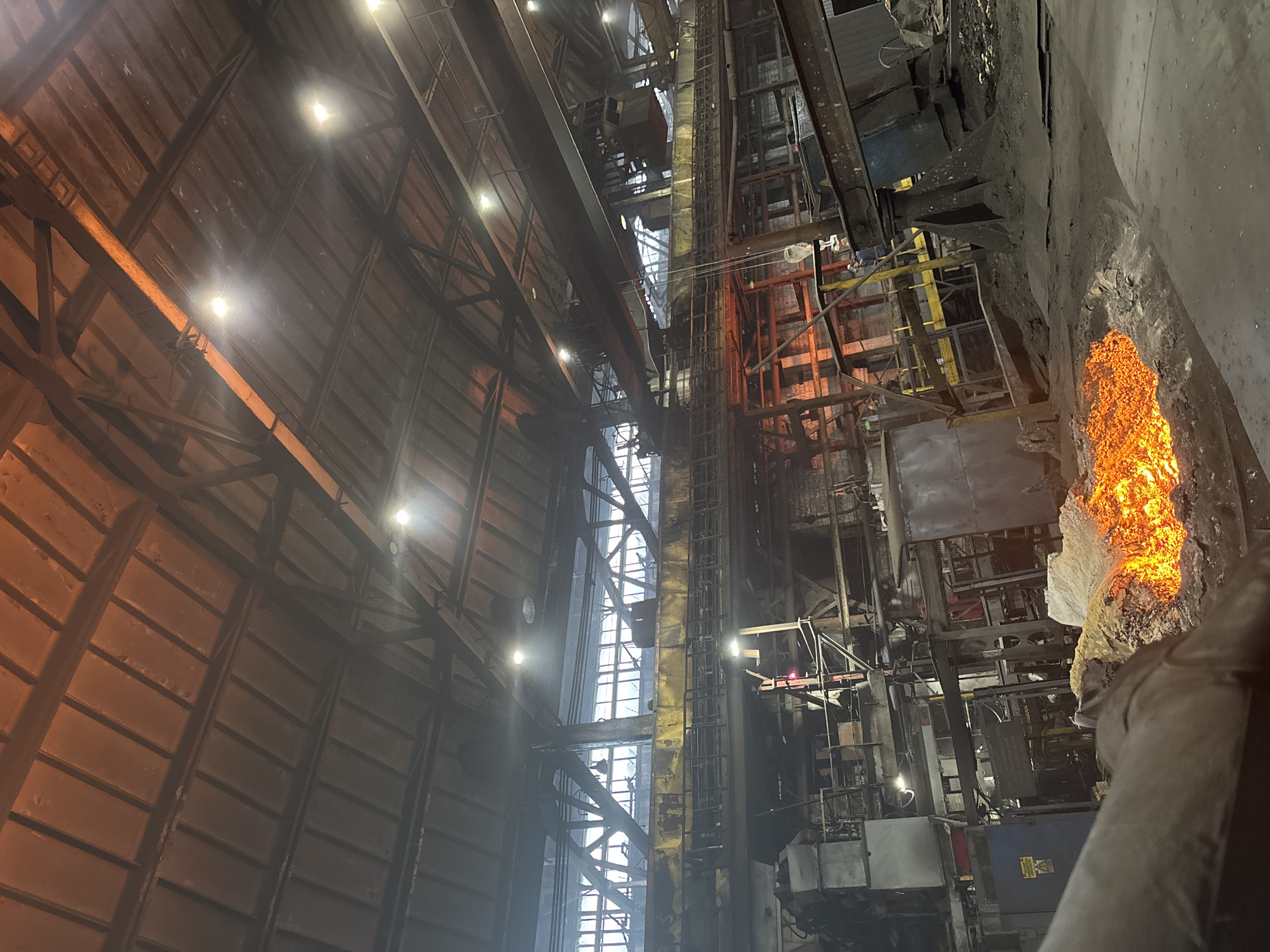 The image depicts the interior of an industrial facility, likely a metallurgical plant, located in the city of Homyel, Belarus. The scene is characterized by a large, cavernous space with a high ceiling supported by exposed metal beams and trusses. Industrial lighting fixtures are visible, casting a warm glow on the metallic structure. In the foreground, a pool of molten material, glowing intensely with orange and yellow hues, sits on the ground, surrounded by debris and industrial equipment. Various structures and machinery are scattered throughout the facility, including metal frameworks, pipes, and what appear to be hoppers or vats. A prominent overhead crane system is visible, with its trolley and hook system positioned above the main industrial floor. The overall environment is gritty and functional, indicative of heavy industrial operations.