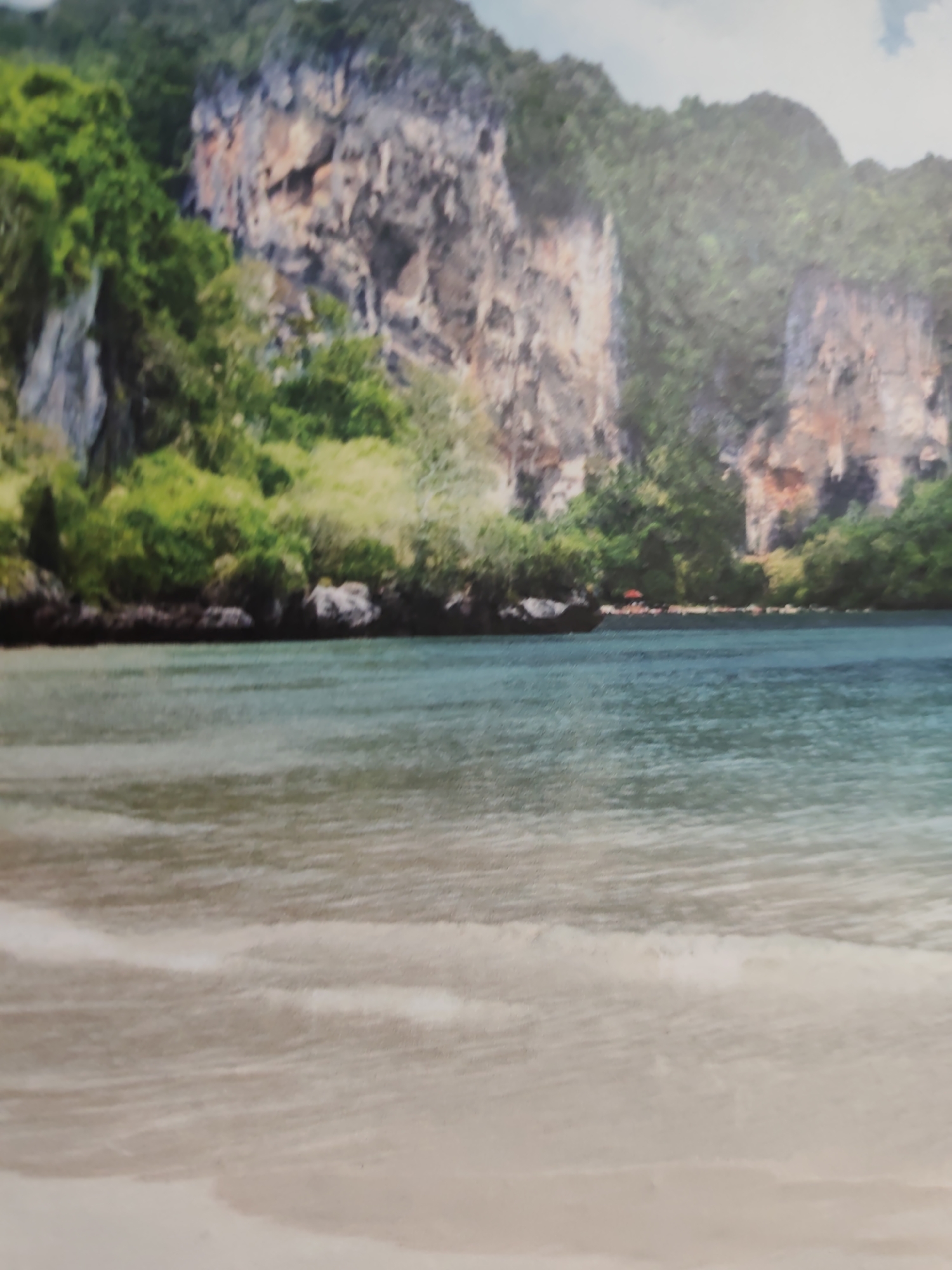 The image depicts a serene tropical coastal landscape. In the foreground, a gentle expanse of light beige sand transitions into clear, shallow turquoise-green water with subtle wave patterns. The midground consists of calm ocean waters reflecting ambient light. The background is dominated by imposing, heavily vegetated limestone cliffs that rise steeply from the water's edge. These karstic formations exhibit varied rock textures and colors, including shades of grey, brown, and reddish-orange, topped with lush green tropical foliage. Along the distant shoreline at the base of the cliffs, dense green trees and shrubs are visible, along with a small red umbrella suggesting a distant beach amenity. No discernible human figures or explicit actions are present. The scene is depicted with a soft-focus aesthetic. This image is located in the city of Pazardzhik, Bulgaria.