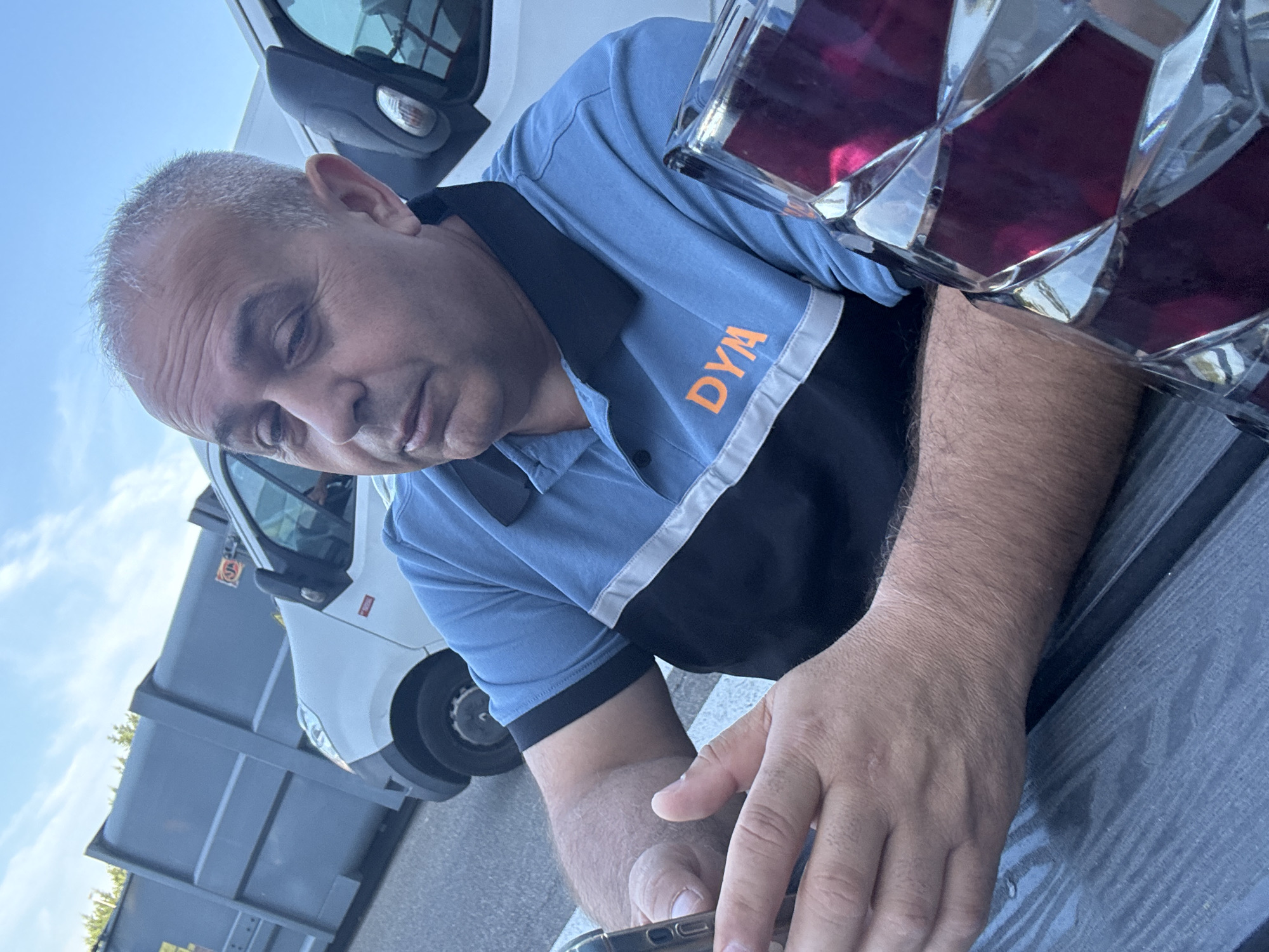 In Boom, Belgium, a middle-aged man with graying hair and olive skin sits at a table, looking down at his smartphone. He is wearing a blue polo shirt with a black collar and a horizontal black and white stripe across the chest, with the orange letters "DYM" visible on the blue portion. His hands are resting on the table as he holds the phone. To his right, a faceted glass object filled with a dark red liquid is partially visible. In the background, a grey dumpster is positioned next to a white van. The scene appears to be outdoors, possibly at a business or industrial location, with a clear sky overhead.