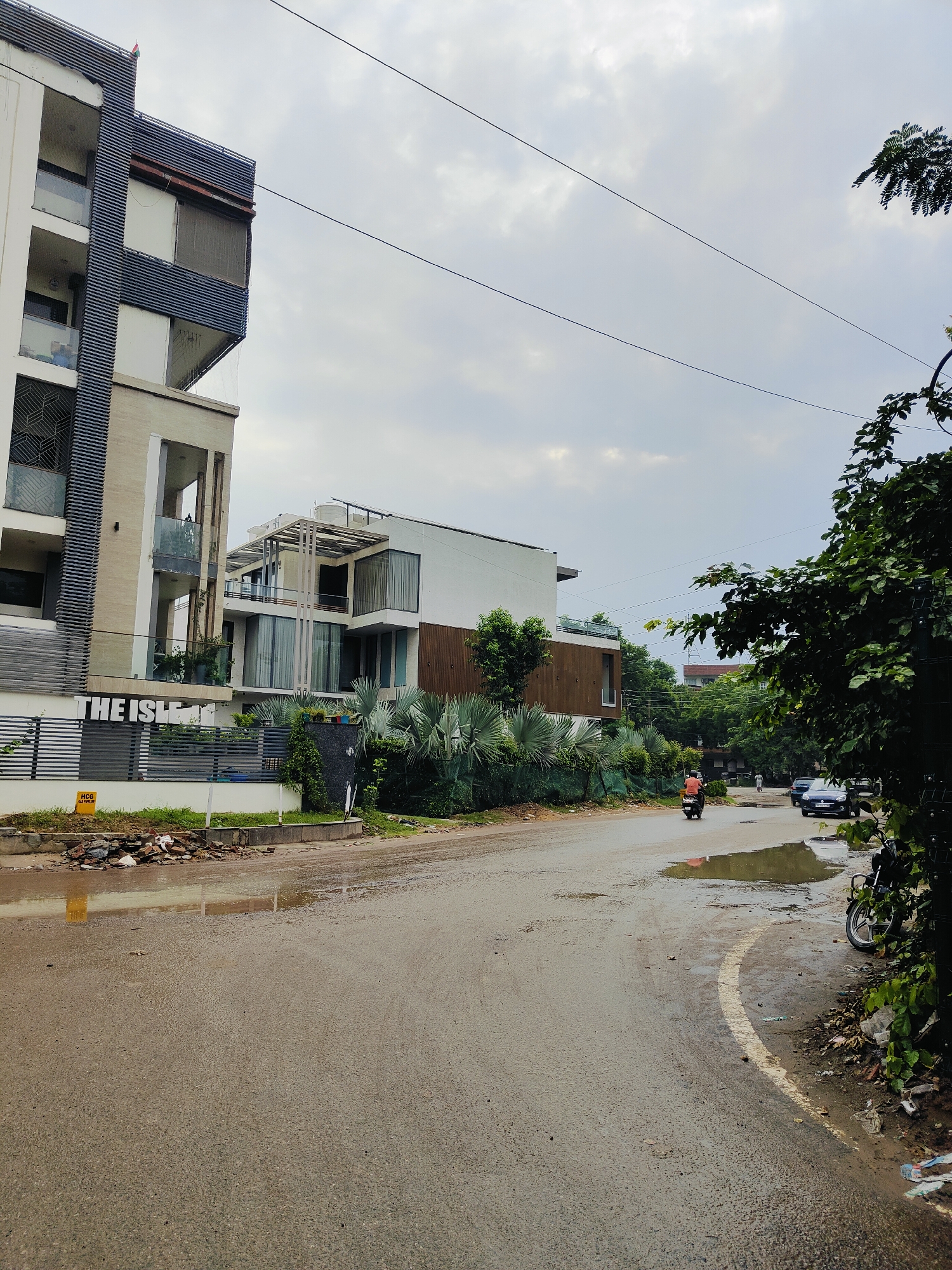 A frontal, medium shot captures a street scene in Gurgaon, India, under an overcast sky. The paved road, wet from rain, curves from the lower right to the mid-left of the frame. Puddles dot the road.
To the left, a modern, multi-story building, "THE ISLE," is visible with balconies and dark, horizontal louvers. A fence runs along the property line, with debris and a yellow sign indicating "HCG GAS PIPELINE" in front of it.
Across the street, another contemporary building features large windows and wooden accents. Lush green foliage, including fan-shaped plants, surrounds the structures.
A person on a scooter travels down the road from left to right. Further down the road, a dark-colored car is parked on the right side, next to lush green trees and undergrowth. A motorcycle is parked in the foreground on the right, partially obscured by foliage. Power lines are visible against the cloudy sky.