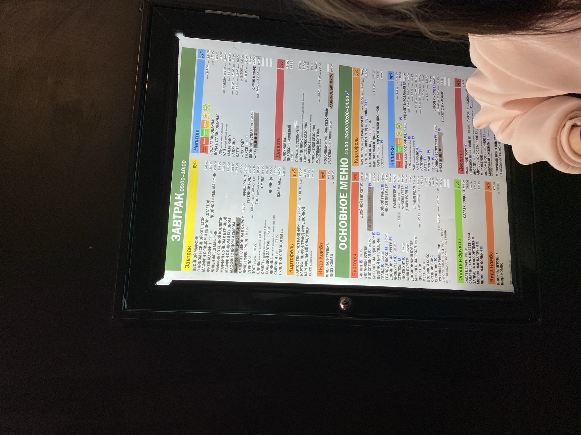 A close-up shot of a lit menu board, encased in a black frame, displays Russian text categorized into "Breakfast," "Drinks," "Desserts," "Burgers," and "Potatoes." The menu details various food items and their prices in rubles. Parts of a person are visible at the right corner of the frame. The image appears to have been taken indoors in a dimly lit environment. The location is Saint Petersburg, Russia.