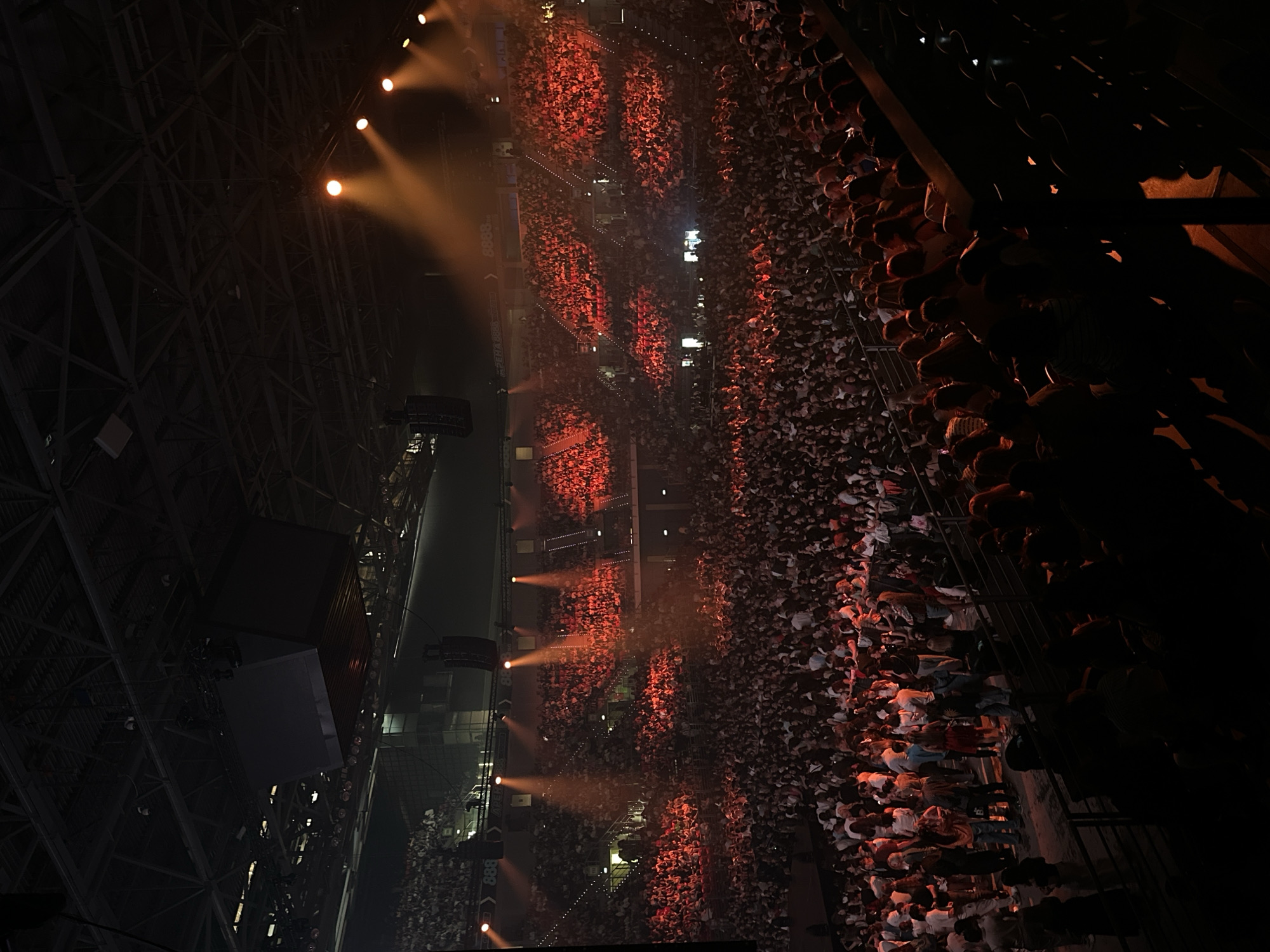 The image shows an elevated view of a large indoor event. The location is likely the Arena Armeec in Sofia, Bulgaria due to the architectural features of the roof and the typical event configuration. A massive crowd fills the arena, primarily dressed in white shirts, and is illuminated by stage lighting creating sections of bright orange. Structural elements of the arena are visible on both the left and right sides, with spotlights positioned along the left. The far right side of the image is partially obscured by darkness.