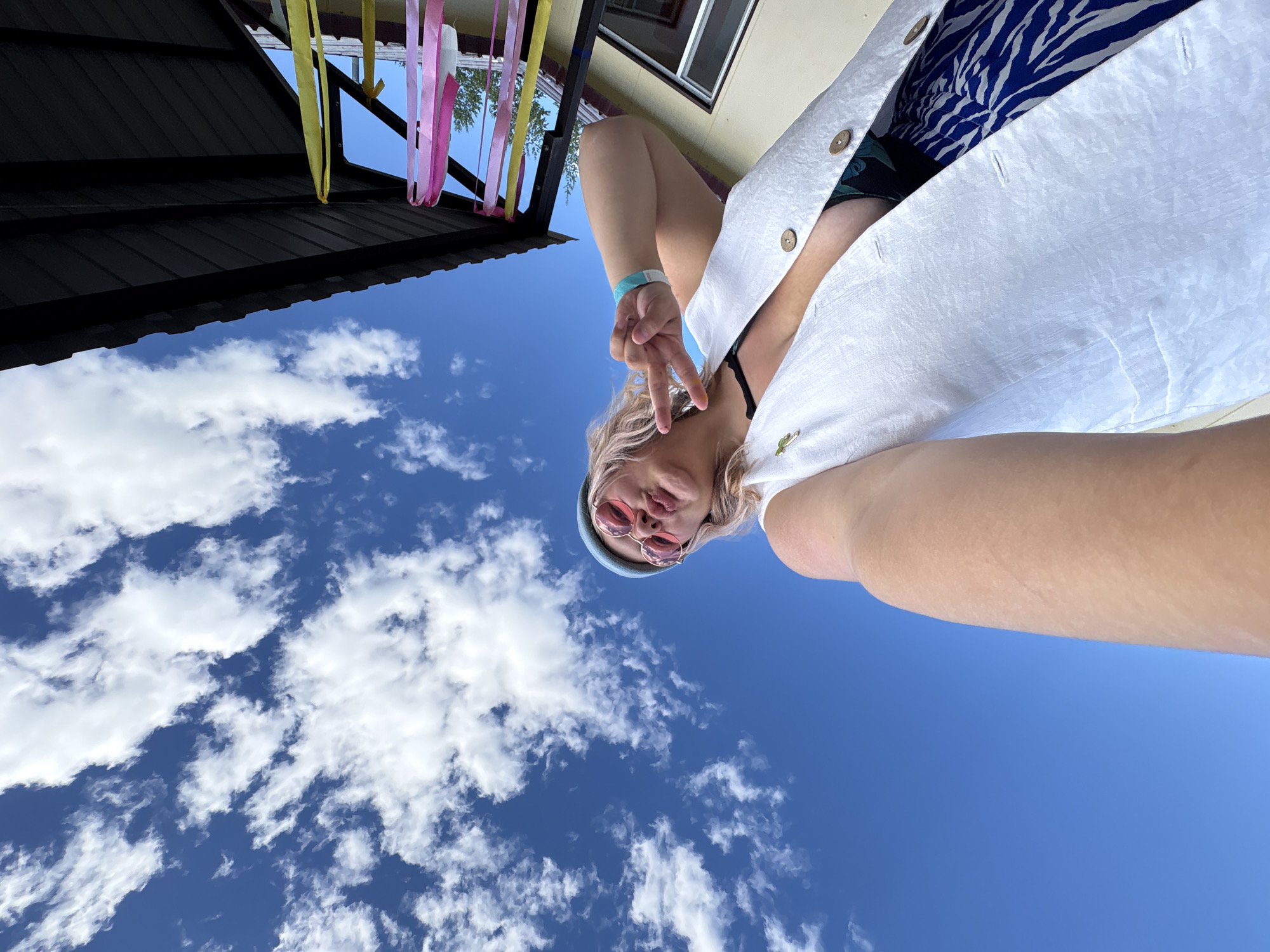 The image shows a woman taking a selfie outdoors. She is wearing a white vest, a blue beanie, and pink-rimmed sunglasses. She is making a peace sign with her right hand. The background consists of a bright blue sky with scattered white clouds, and part of a dark-colored roof with pink and yellow streamers hanging down. The lower part of the image shows the woman's arm and torso, and a blue and white patterned skirt. There is no information in the image to suggest the location is in Birobidzhan, Russia.