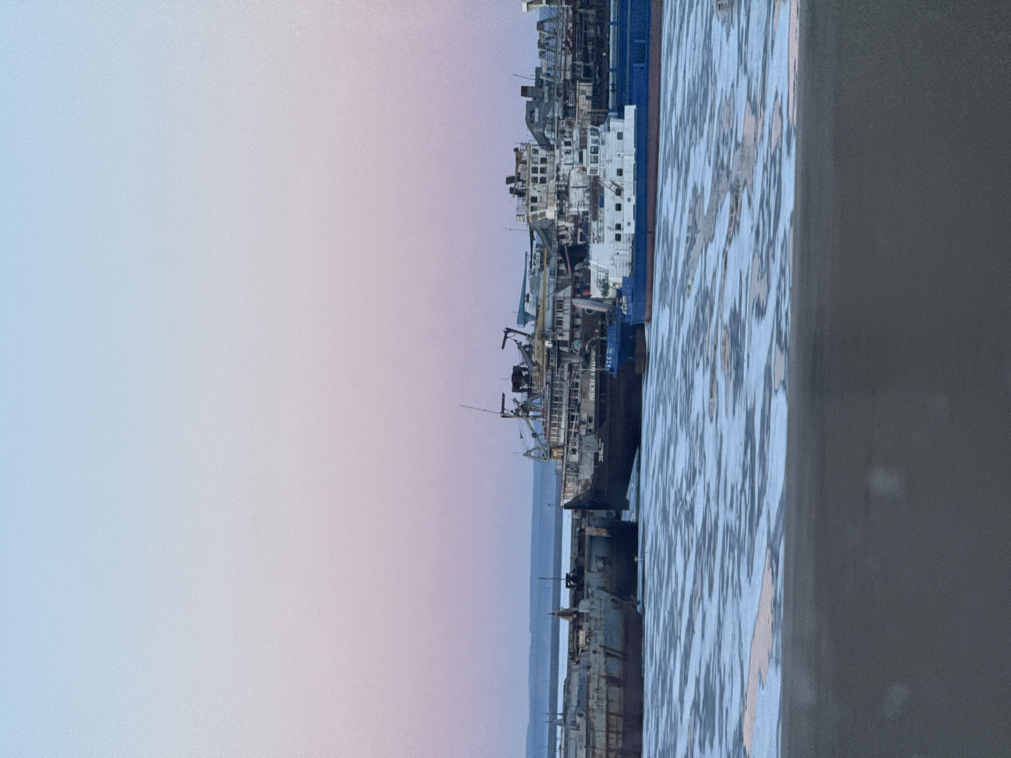 The image shows a view of a shipyard in the city of Chaikovsky, Russia, with numerous vessels docked alongside a concrete pier. The sky is a gradient of light blue to pale purple. The ships vary in size and type, with some appearing older and rusted, while others are newer and painted white and blue. The water is rippled, reflecting the sky and the ships. A gray vertical structure is visible on the right edge of the image.