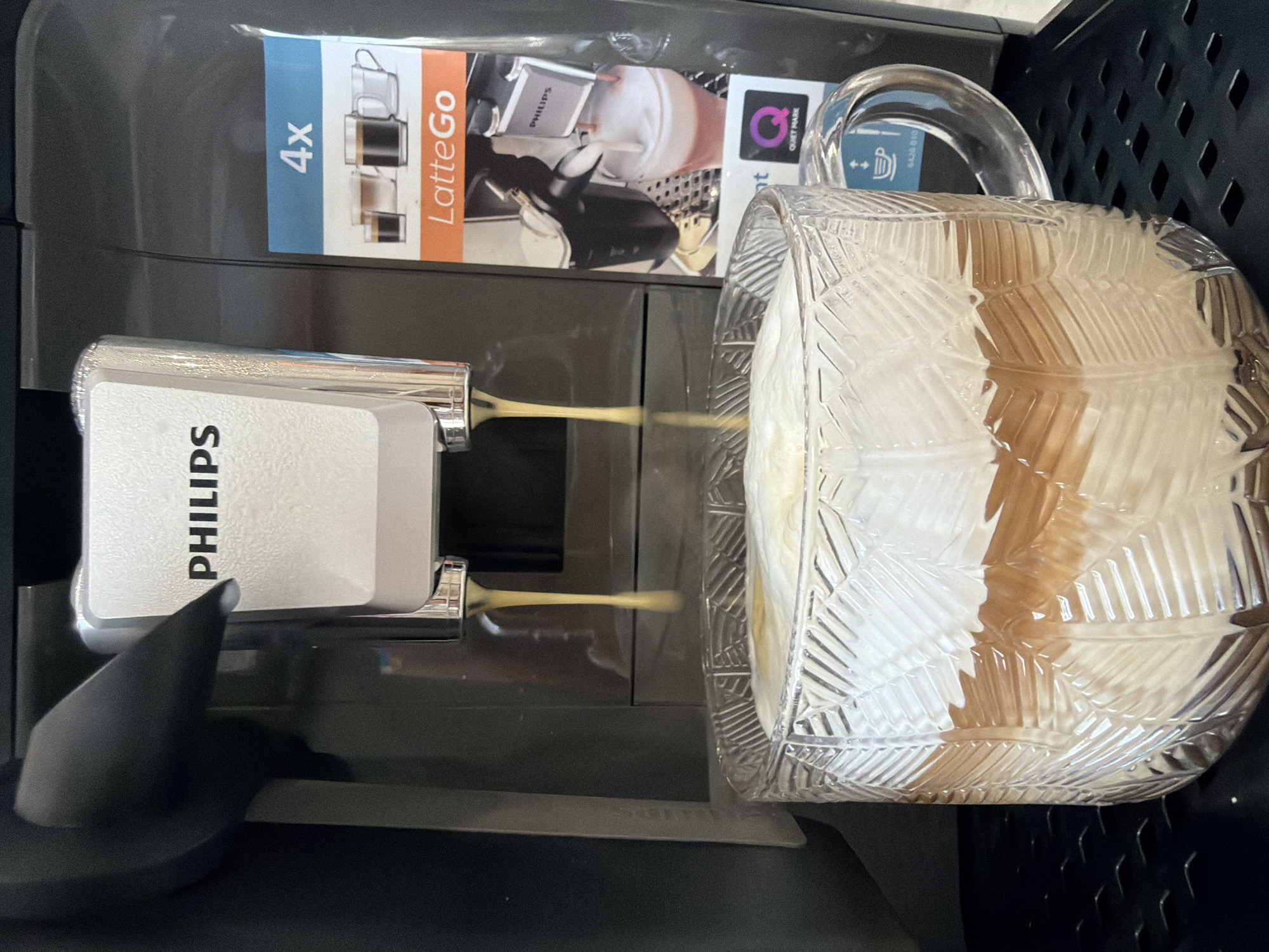 A Philips coffee machine, located in Chaikovsky, Russia, is actively dispensing a light brown liquid from two nozzles into a clear, textured glass mug. The machine is primarily dark gray/black, with a prominent metallic silver dispenser unit marked "PHILIPS." A vertical label on the machine's right side displays "4x," "LatteGo," illustrations of various coffee cups, a Philips milk frother, and a "Quiet Mark" logo. The clear glass mug, featuring a patterned exterior, contains a layered beverage with a substantial white foam top layer over a light brown liquid. The mug rests on a black, textured drip tray. The background is out of focus.