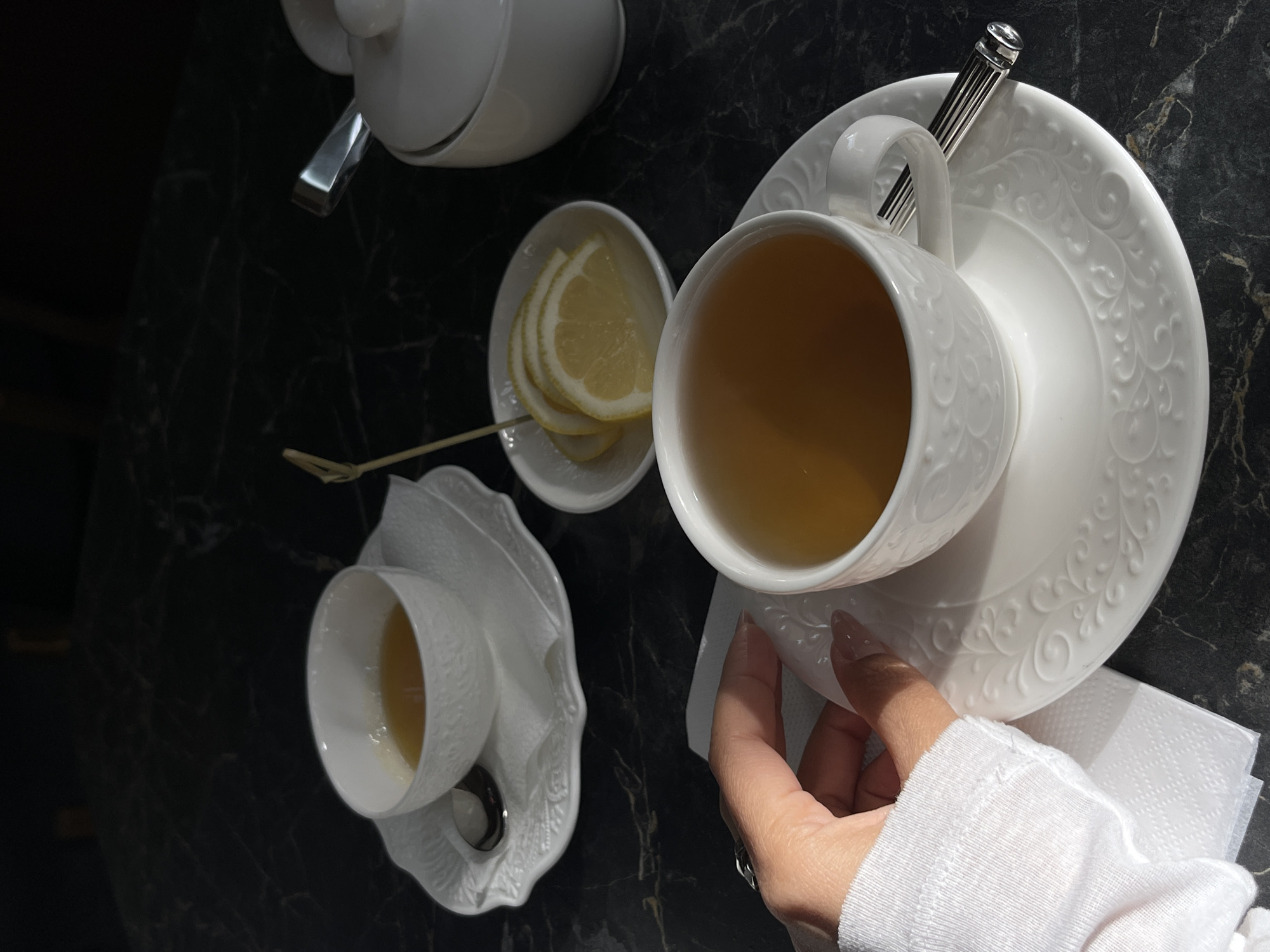 The image presents an overhead view of a tea service arranged on a dark, possibly marble or granite, surface. The scene features several key elements: a white porcelain teapot, a small dish containing lemon slices, and two cups and saucers filled with tea. A silver teaspoon rests on one of the saucers.
A person's hand, adorned with a ring and wearing a white long-sleeved shirt, is partially visible. The hand appears to be holding a stack of white napkins or small cloths. The light in the photo suggests it could be taken during the daytime, perhaps by a window or in a well-lit indoor area.
Without additional details, pinpointing the exact location in Sochi, Russia, is difficult. However, based on the objects and atmosphere, it could plausibly be a cafe, restaurant, or hotel where high tea or a similar service is offered.