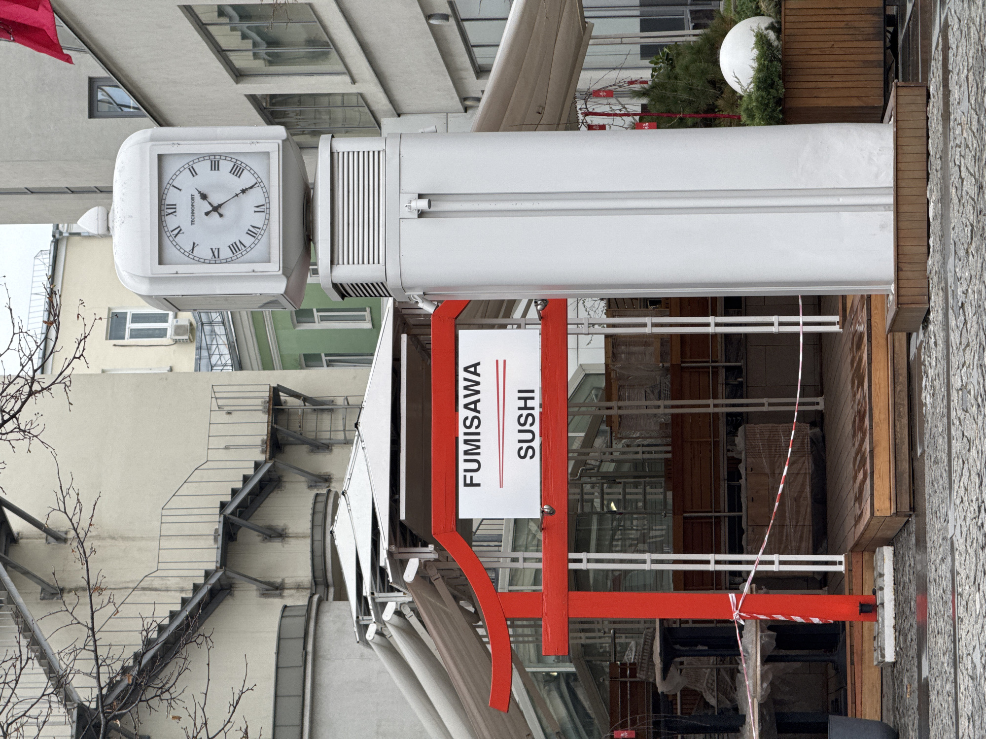 This media file captures an urban street scene in Moscow, Russia, during what appears to be an overcast day. The image is taken from a slightly low angle, looking towards several buildings and a commercial establishment.
In the left foreground, a prominent white square-faced clock is affixed to a tall, minimalist white pillar. The clock displays Roman numerals and reads "TECHNOPORT" below its center. The clock hands indicate the time is approximately 1:55. Below the clock, a white canopy or awning extends over a commercial space.
To the right of the clock and under the canopy, a sign for a sushi restaurant is clearly visible, reading "FUMISAWA SUSHI" in red and white lettering. The restaurant's frontage features striking red architectural accents, including a curved element and vertical supports. Below the sign and red structures, white barricades, red and white striped caution tape, and wrapped construction materials suggest that the area is either undergoing renovation or is temporarily closed.
The background consists of several multi-story buildings with light-colored facades and various window styles. One building in the mid-ground features a visible external metal staircase. Bare tree branches are visible in the far left and right foreground, suggesting a late autumn or winter season. To the right of the main pillar, a patch of greenery with small trees and a large white spherical outdoor light fixture adds a touch of landscaping. The ground in the foreground and to the far right is paved with textured stones or cobblestones. No people are visible in the scene.