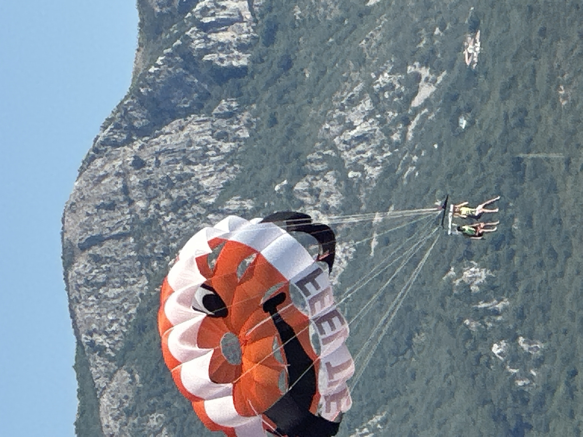 In Boreti, Montenegro, two individuals are parasailing against a backdrop of a rugged, vegetated mountain under a clear blue sky. A brightly colored parasail, predominantly orange and white with black accents and a graphic design resembling a face, is prominently featured in the foreground. The harness below the parasail carries two people, one wearing a green top and the other a yellow top, both with legs dangling. The mountainous terrain behind them is characterized by rocky outcrops and dense green foliage, with a few scattered buildings visible in the distance on the lower slopes.