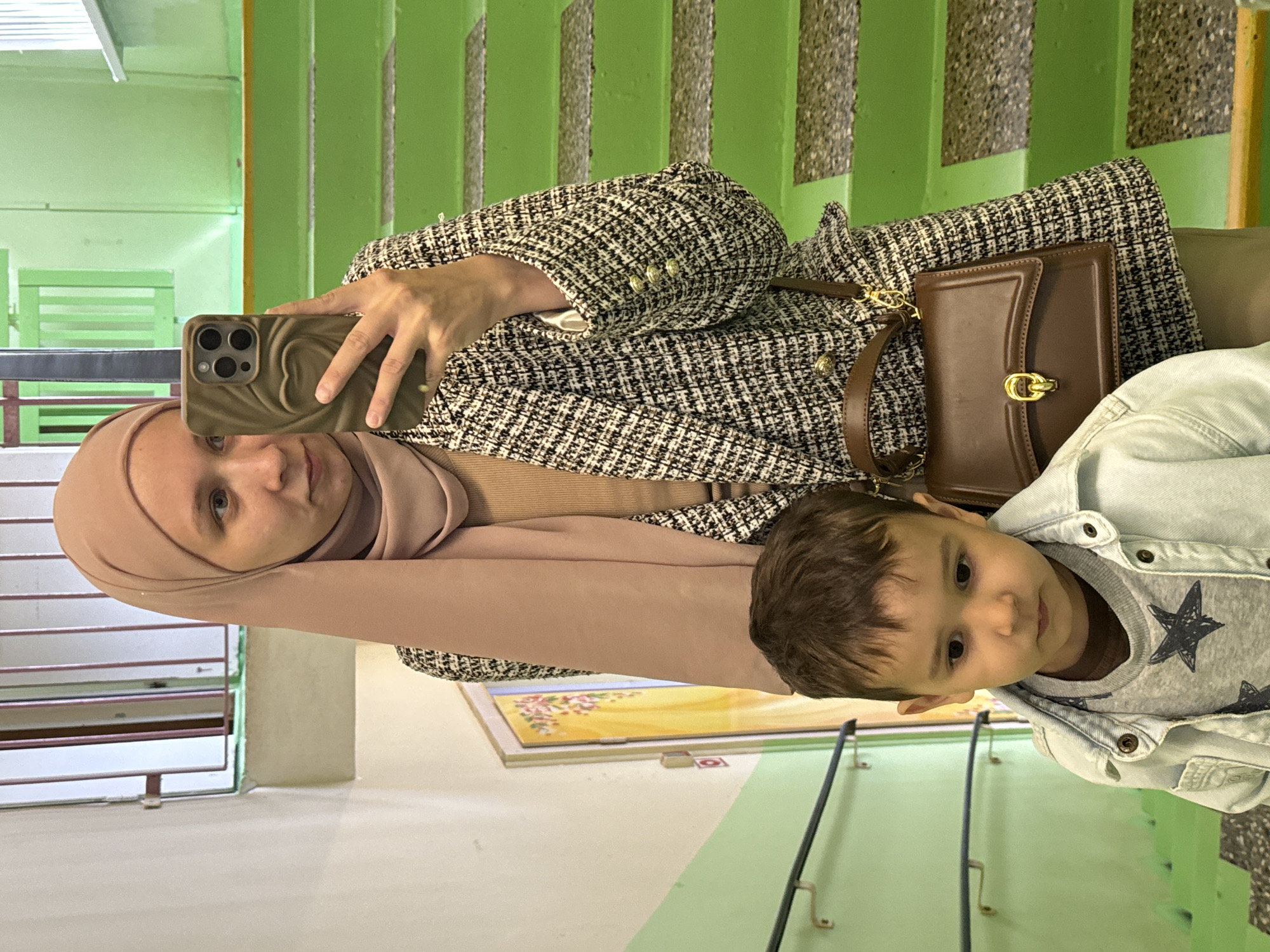 A woman wearing a hijab and a tweed jacket is taking a selfie with a young child in front of a green staircase. The woman is holding a smartphone with a brown case. She is wearing a beige hijab and a tweed jacket with gold buttons. She has a brown leather purse with a gold clasp. The child is wearing a grey shirt with black stars and a denim jacket. They are standing on a landing with a picture hanging on the wall. The staircase is painted green. The location is Chaikovsky, Russia.