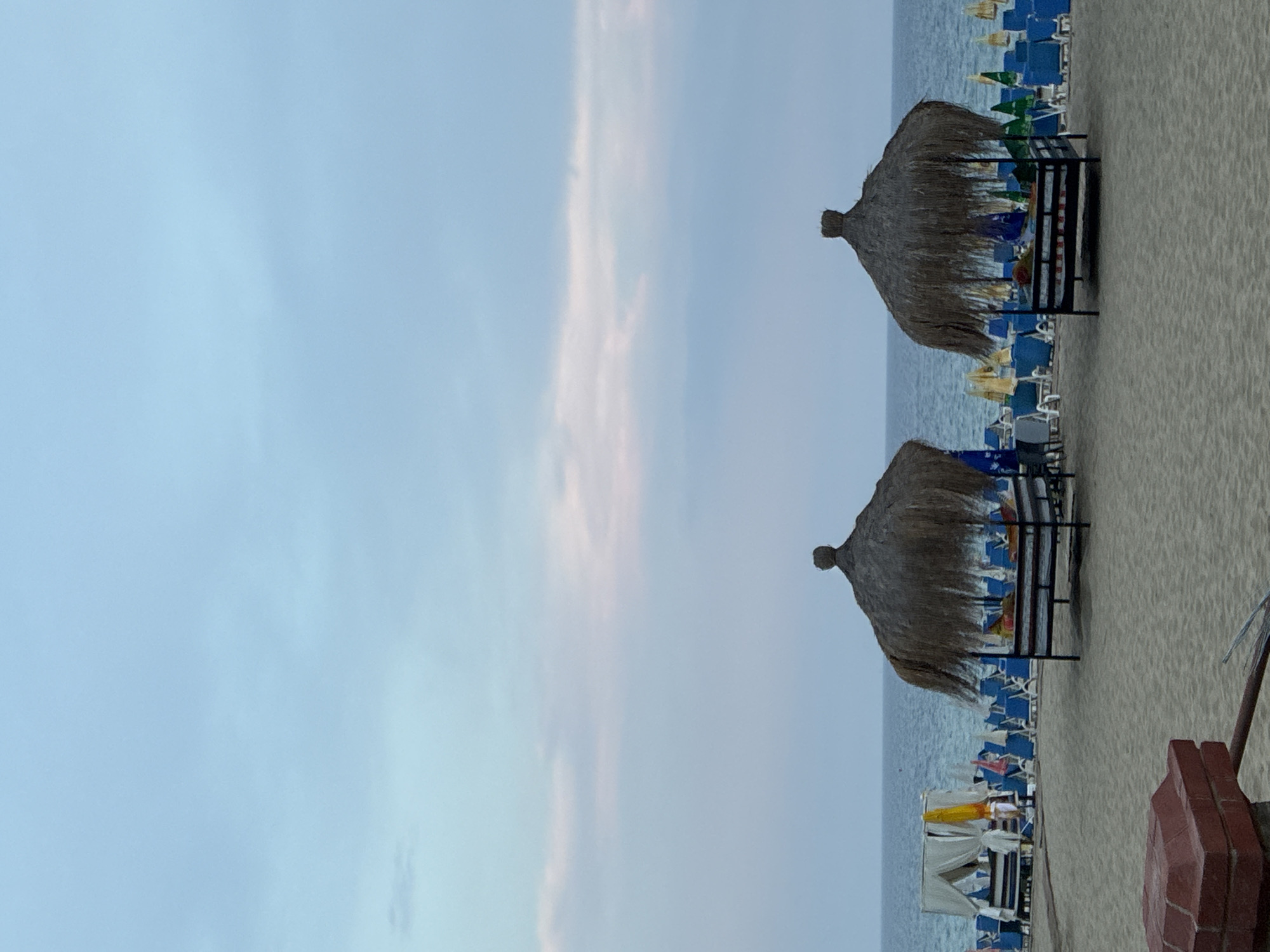 The image depicts a beach scene in Alanya, Turkey, likely during late afternoon or early evening due to the soft lighting and pastel hues in the sky. Two thatched cabanas, elevated on dark wooden platforms, are prominently featured in the foreground, offering shaded seating areas. Each cabana contains cushions and appears to be unoccupied.
Behind the cabanas, rows of empty blue beach chairs and loungers stretch towards the sea, interspersed with yellow and green beach umbrellas. In the middle ground, a white tent-like structure with draped curtains is set up, also appearing unoccupied. The sea is a calm, deep blue, meeting the horizon under a pale sky painted with wisps of pink and white clouds. The beach itself is sandy and appears well-maintained, with a low, red brick structure visible in the bottom left corner. The overall atmosphere is peaceful and suggests a resort setting preparing for or winding down after a day at the beach.