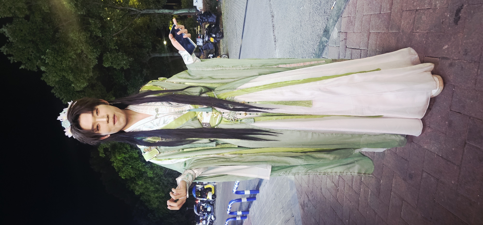 A person in traditional Chinese attire stands on a brick sidewalk at night in the Chongqing High-tech Zone, China. The individual wears a long, flowing light green and white robe with elaborate embellishments, including gold trim, tassels, and what appears to be a floral crown on their head. Their long, dark hair cascades down their back and shoulders. The person holds a dark-colored smartphone in their right hand and has their left hand extended outwards. In the background, trees, a street with vehicles, and streetlights are visible, indicating an urban outdoor setting. The lighting suggests it is nighttime.