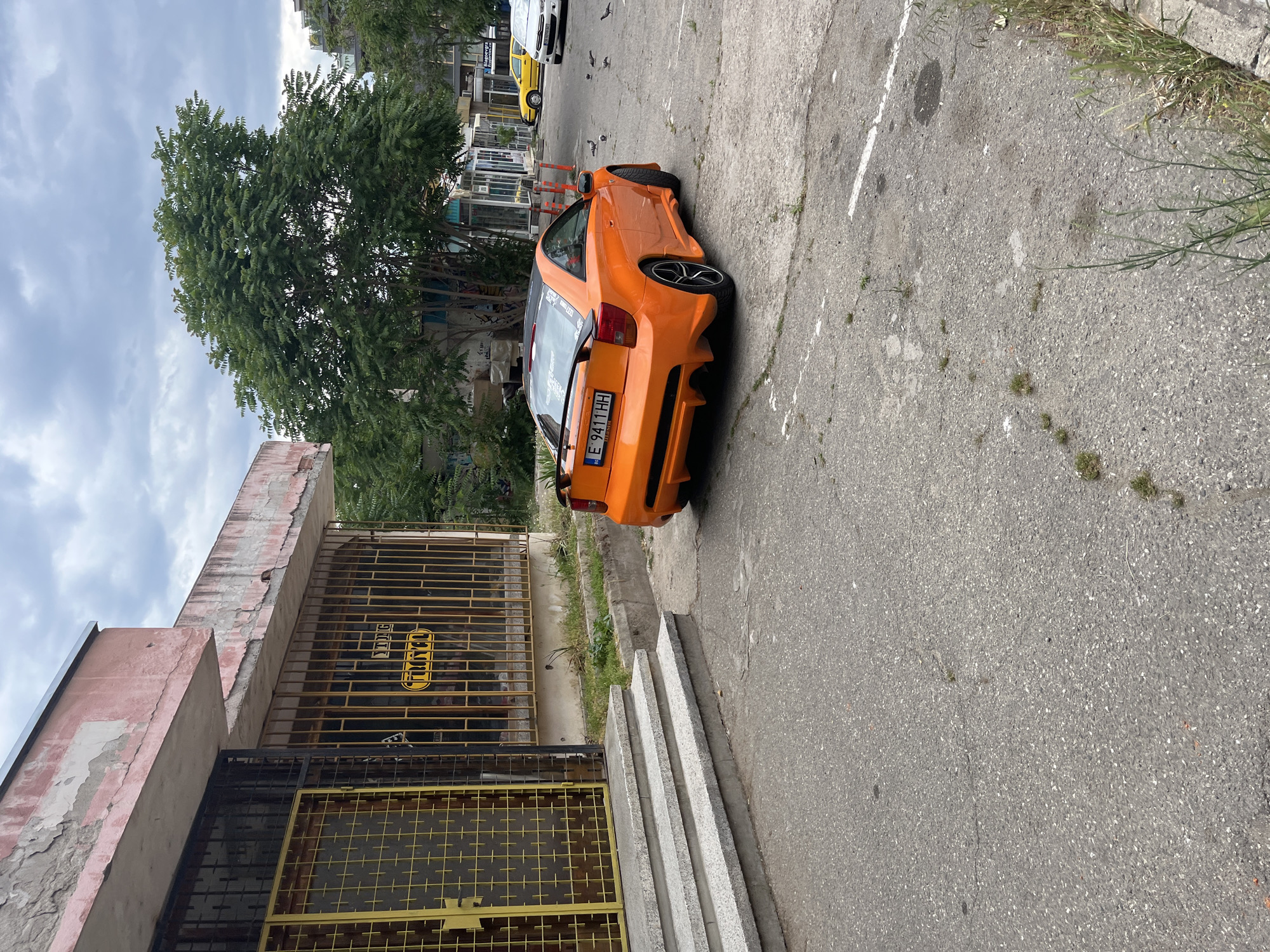 The image depicts an orange Toyota Celica parked on a grey asphalt surface in Blagoevgrad, Bulgaria. The car is positioned near a building with a weathered pink facade and a grated metal gate. A tree with dense green foliage is visible in the background. Other vehicles are parked further down the street. The sky is cloudy. A concrete curb and several steps are situated between the building and the road.