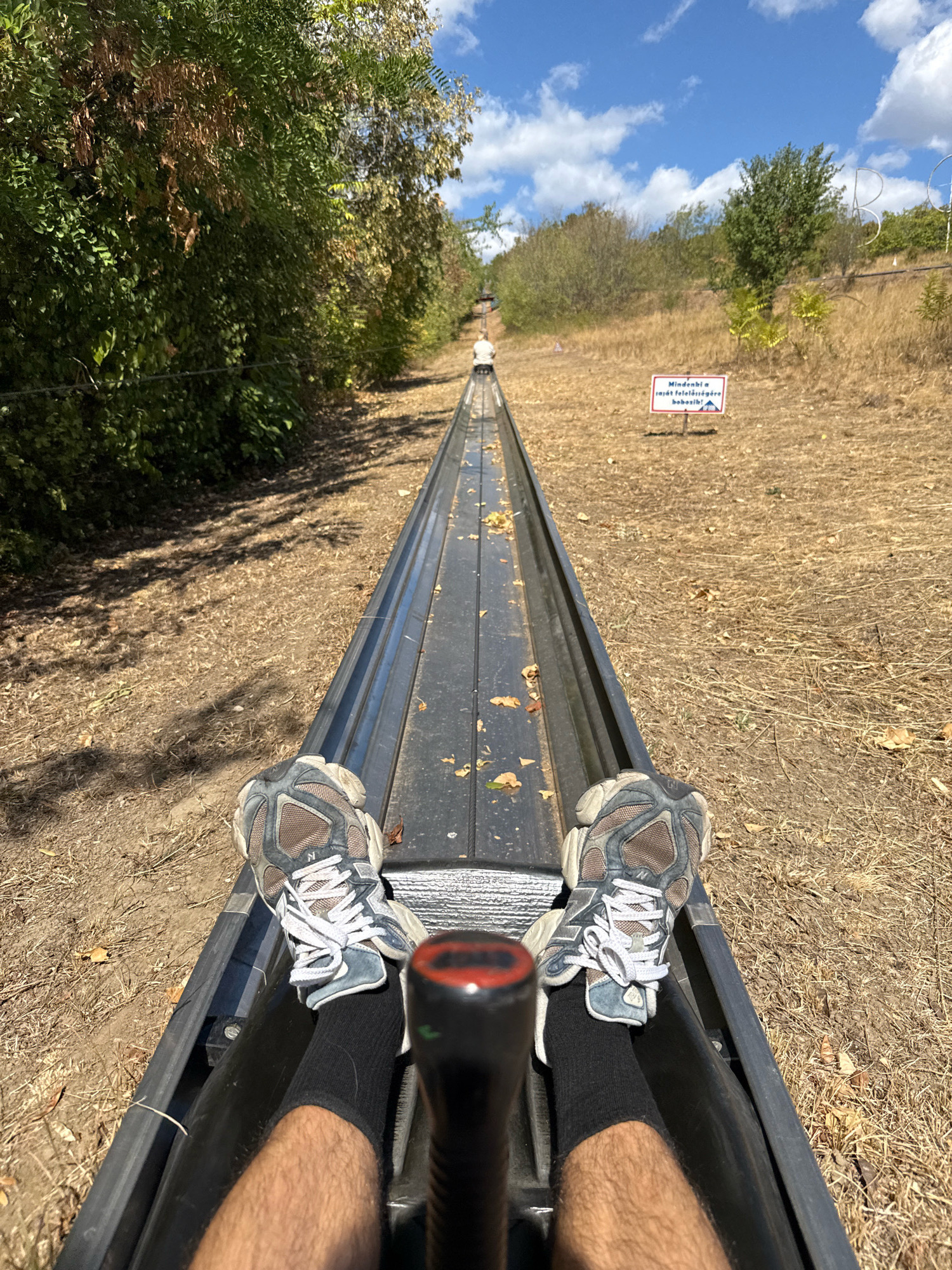 The image captures a first-person perspective of someone seated in a bobsled-like vehicle on a gravity-powered roller coaster. The vehicle is on a track made of dark metal, with fallen leaves scattered on it. The user's legs and feet, clad in athletic shoes and black socks, are prominently visible at the front of the vehicle. The track winds downhill through a dry, grassy landscape with sparse trees and bushes on either side. In the background, another person is visible further up the track, also in a bobsled. A sign with Hungarian text and an image of a person on a sled is posted on the right side of the track. The sky is blue with scattered white clouds. This scene is located at the Memento Park's Bobsled Track in Budapest, Hungary, a popular tourist attraction offering scenic views and a thrilling ride.