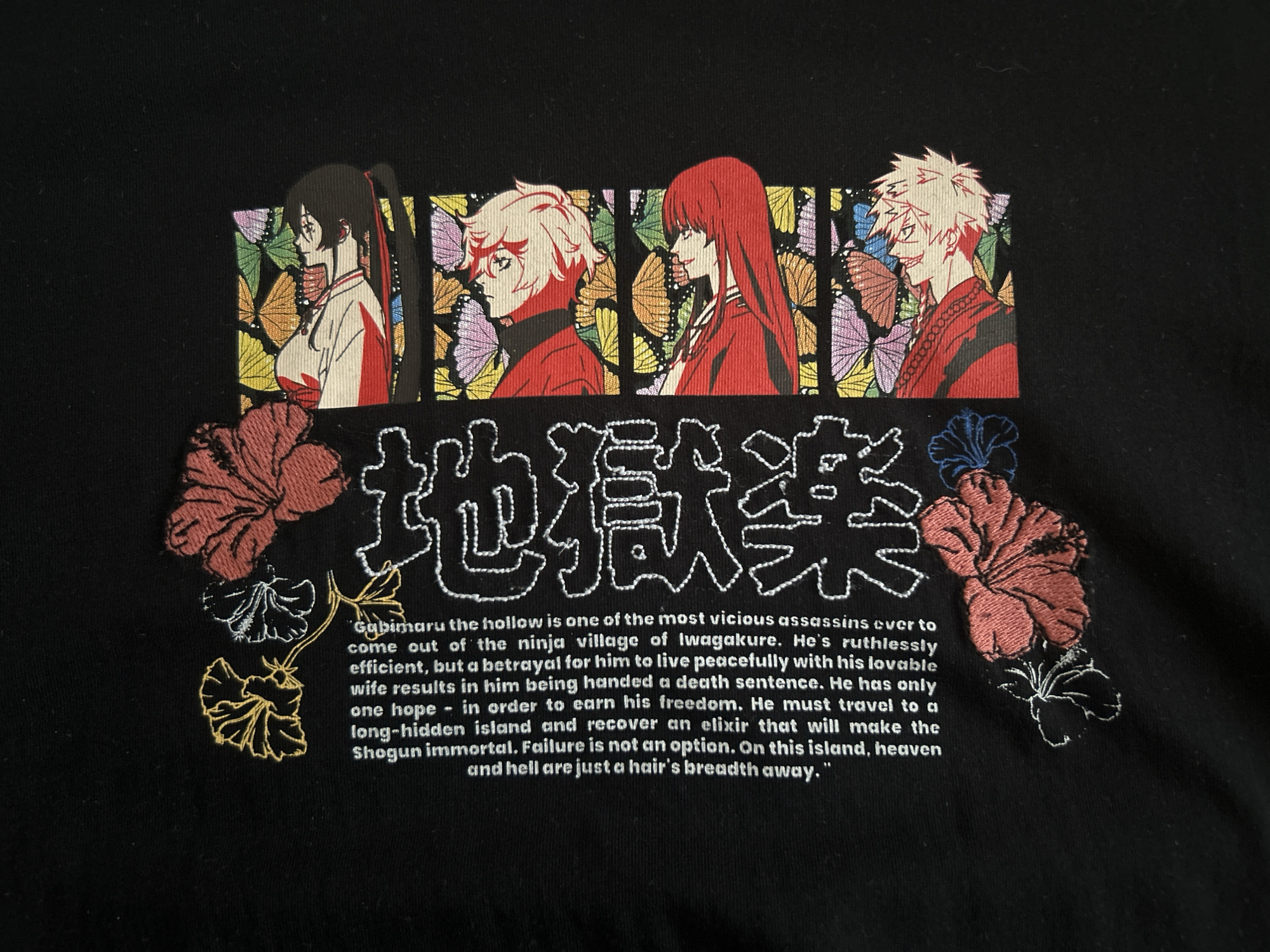 The image shows a black t-shirt with an embroidered design. The design consists of four anime-style character portraits in profile, set against a backdrop of stylized butterflies. Below the portraits is a line of Japanese characters embroidered in white. Underneath the Japanese characters is a block of English text, also embroidered in white, describing the character "Gabimaru the hollow." The text is flanked by embroidered hibiscus flowers in red, white, yellow, and blue. The shirt appears to be photographed indoors on a flat surface.