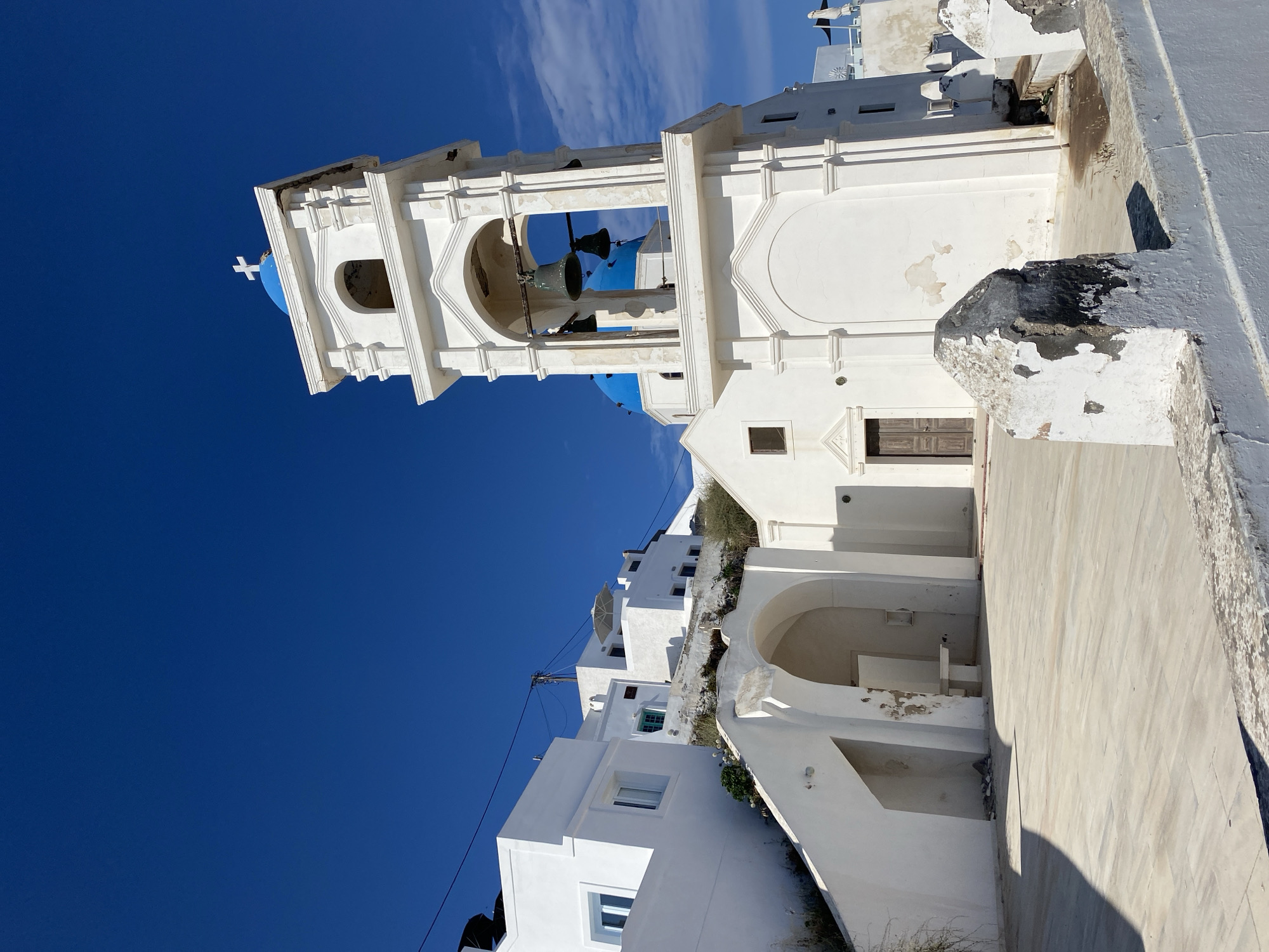 This is a vertical, eye-level, outdoor, medium shot of a white church with blue domes against a bright blue sky with some wispy white clouds. The church has a bell tower with arched openings containing bells. A white cross tops a blue dome. The architecture is traditional of Santorini, Greece, with whitewashed buildings. The image is well-lit by the sun, indicating a clear, sunny day, likely in the morning or afternoon due to the shadows. The scene appears to be a tourist destination, likely in a village on Santorini, given the iconic architecture. There is no visible text in the image.
