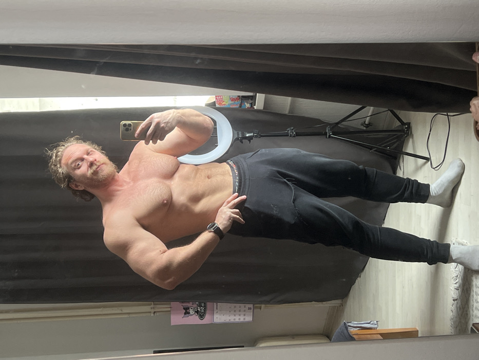 An adult male stands shirtless in an indoor environment within Saint Petersburg, Russia, capturing a mirror selfie with a light-colored smartphone held in his left hand. He exhibits a muscular physique, with visible development in his chest, shoulders, and arms. His hair is curly and light-colored, and he has a reddish-blonde beard and mustache. A dark wristwatch is present on his left wrist. He is wearing black sweatpants with visible wear and light grey/white ankle socks. His right hand is placed on his hip as he poses.
The immediate background, reflected in the mirror, includes dark brown curtains on both sides of the frame. To the man's right, a white illuminated ring light on a black tripod stand is visible. Beyond the ring light, a window sill holds several small, indistinct objects. To the man's left, a pink wall calendar featuring an anime-style character is affixed to a light-colored wall. The floor is light-colored wood, with a small, light-colored rug partially visible in the bottom-left foreground. A dark cable is coiled on the floor near the ring light. The scene appears to be a casual room, likely an apartment or private residence.