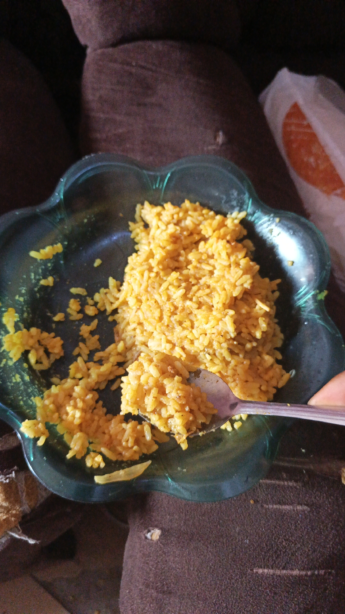 A partially eaten bowl of yellow rice sits on a brown textured surface, possibly a couch or armchair. The rice is in a flower-shaped, translucent green bowl. A silver spoon holds a portion of rice. Part of a white plastic bag with orange markings is visible in the background. The location is unknown.