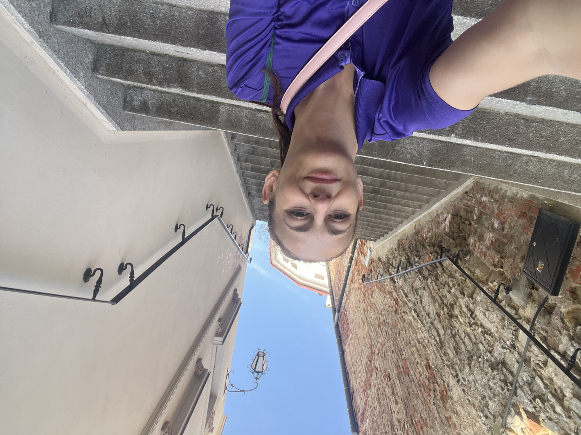 A woman is taking a selfie on a set of outdoor stairs. She is wearing a purple collared shirt and has a pink strap across her shoulder. The stairs are made of stone and are flanked by a brick wall on the left and a light-colored building on the right. There is a metal railing running along both sides of the stairs. Above, a street lamp is visible against a clear blue sky.