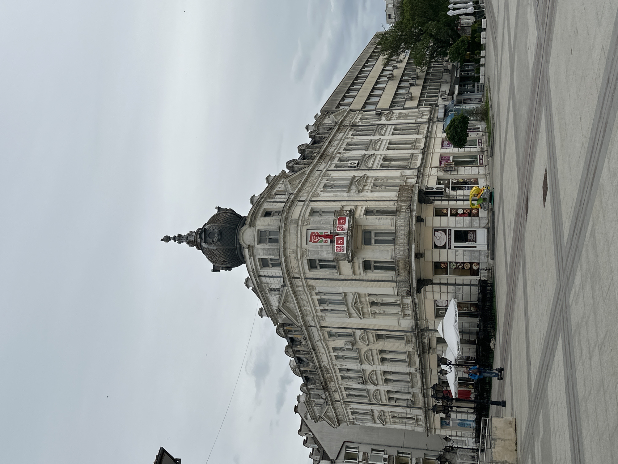 The image shows a view of the Krastata Kazarma building in Vidin, Bulgaria. It is a historic building with a distinctive dome and ornate facade. The building is located on a pedestrian square with light grey stone paving. There is a tall building directly attached to the right with many windows and air conditioning units hanging from the windows. A few people are visible on the edge of the frame, some sitting at tables under large umbrellas. A small merry-go-round animal ride is present. The sky is overcast.