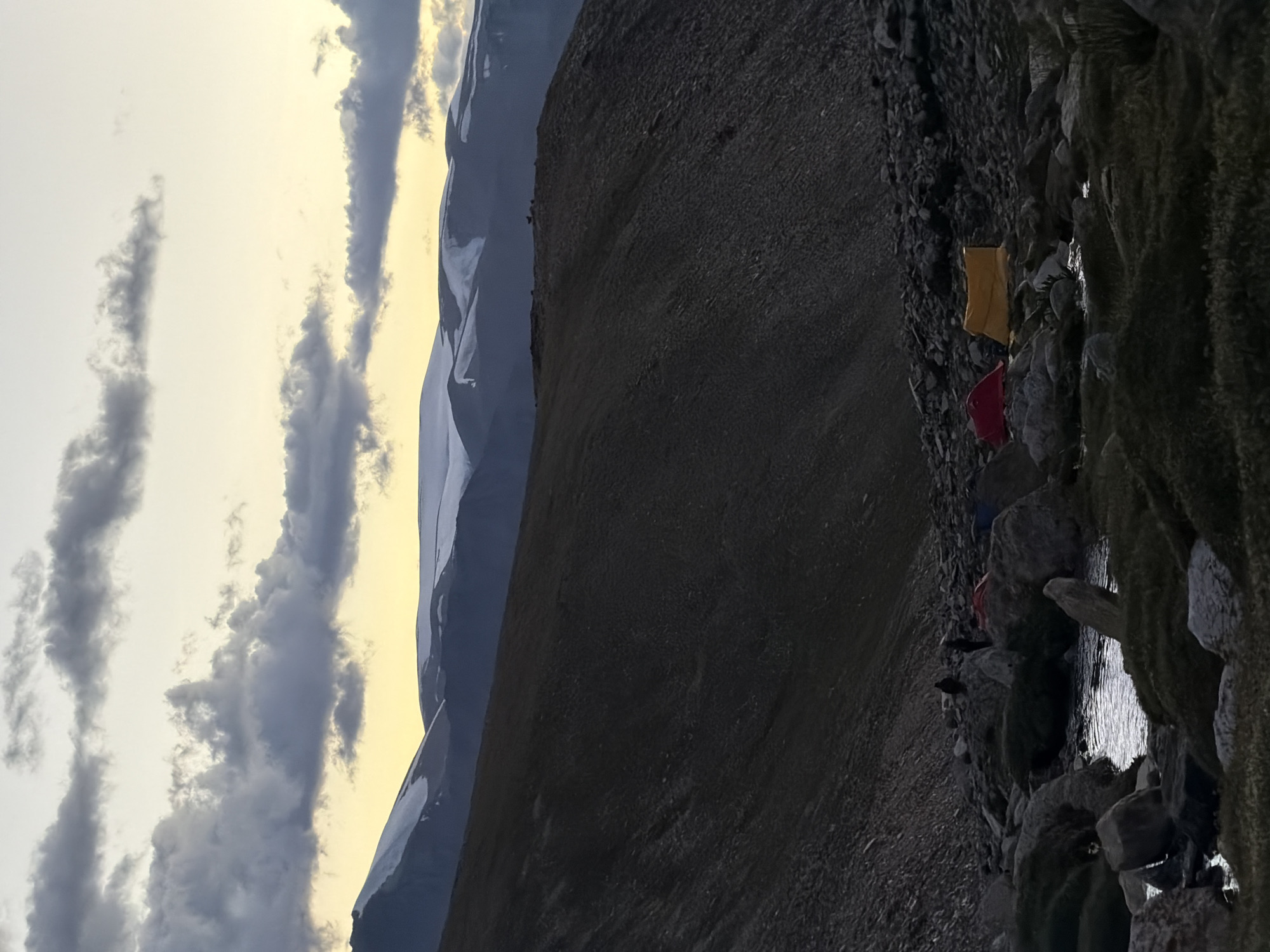 A high-angle, outdoor, medium shot captures a rocky, mountainous landscape at dawn or dusk in an unspecified Indian location. The sky is a pale yellow, streaked with dark, heavy clouds. In the distance, snow-capped mountains form a jagged horizon. The foreground features a dark, scree-covered slope leading down to a rocky stream bed with patches of green vegetation. Two small tents, one red and one yellow, are pitched near the stream. Several figures, appearing as dark silhouettes, are scattered among the rocks and near the tents. In the mid-ground, a ridge of the mountain is visible, with a few distant figures or animals silhouetted on its crest. The overall atmosphere is one of rugged wilderness and transient human presence.