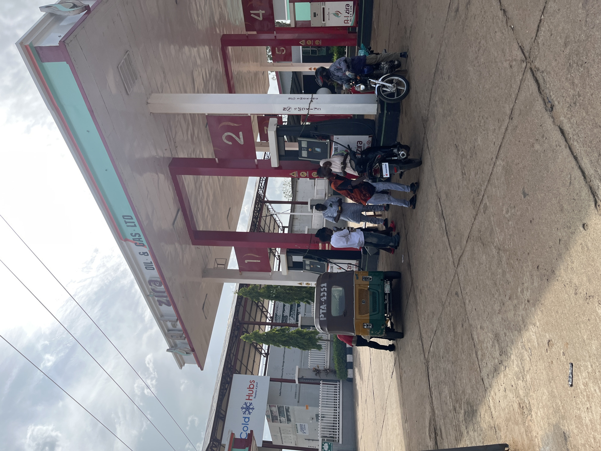 This image captures a scene at a gas station in Jos, Nigeria, likely during the daytime under an overcast sky. The main subjects are several people and various vehicles, including motorcycles and a three-wheeled auto-rickshaw. The gas station has a prominent red and white canopy with fuel pumps visible.
One person is on a motorcycle, appearing to be interacting with the pump or another individual. Further down, a group of people are gathered around two motorcycles. One person is seated on a motorcycle, while others stand nearby, suggesting a moment of rest, conversation, or waiting. A three-wheeled auto-rickshaw, primarily green and yellow, is parked prominently in the foreground. It has the license plate "PTA-4351" visible.
Visible text includes branding for "zira OIL & GAS LTD" on the gas station's canopy and a sign for "Cold Hubs" which reads "Keeping & fresh". There are also smaller signs with "NO SMOKING" and "WELCOME TO" printed on them. The overall impression is a busy, everyday scene at a local fuel station. The lighting suggests it's daytime, and the clouds indicate potentially mixed or overcast weather. The ground is paved with concrete.