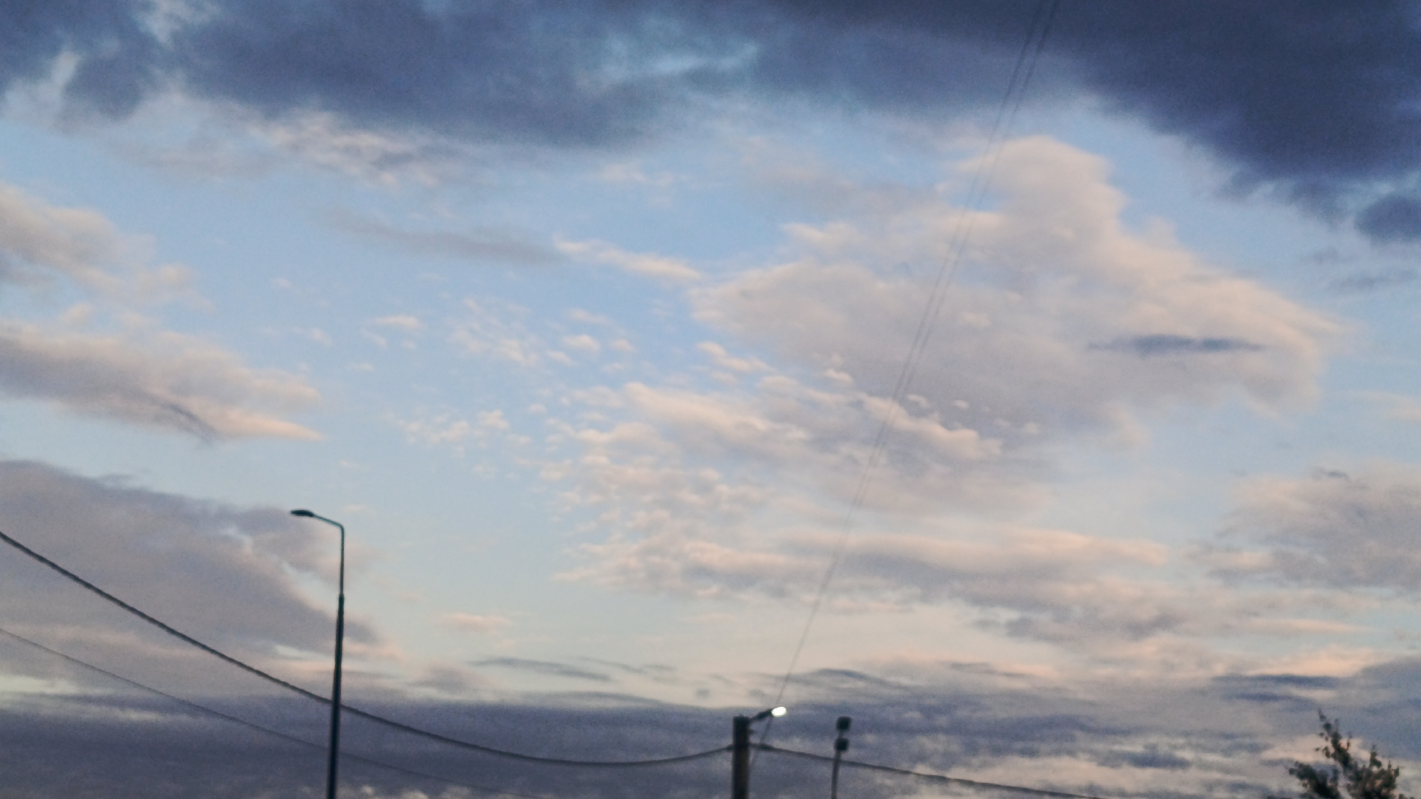 The image shows a cloudy sky with a streetlight and power lines in the foreground. The sky is a mix of blue, white, and gray clouds, suggesting it is either early morning or late evening. The streetlight is a modern design with a curved arm. The power lines are strung across the frame, indicating an urban or suburban setting. The scene appears to be outdoors, likely in a street or park. There are no people visible in the image. The location is Zvenigorod, Russia.