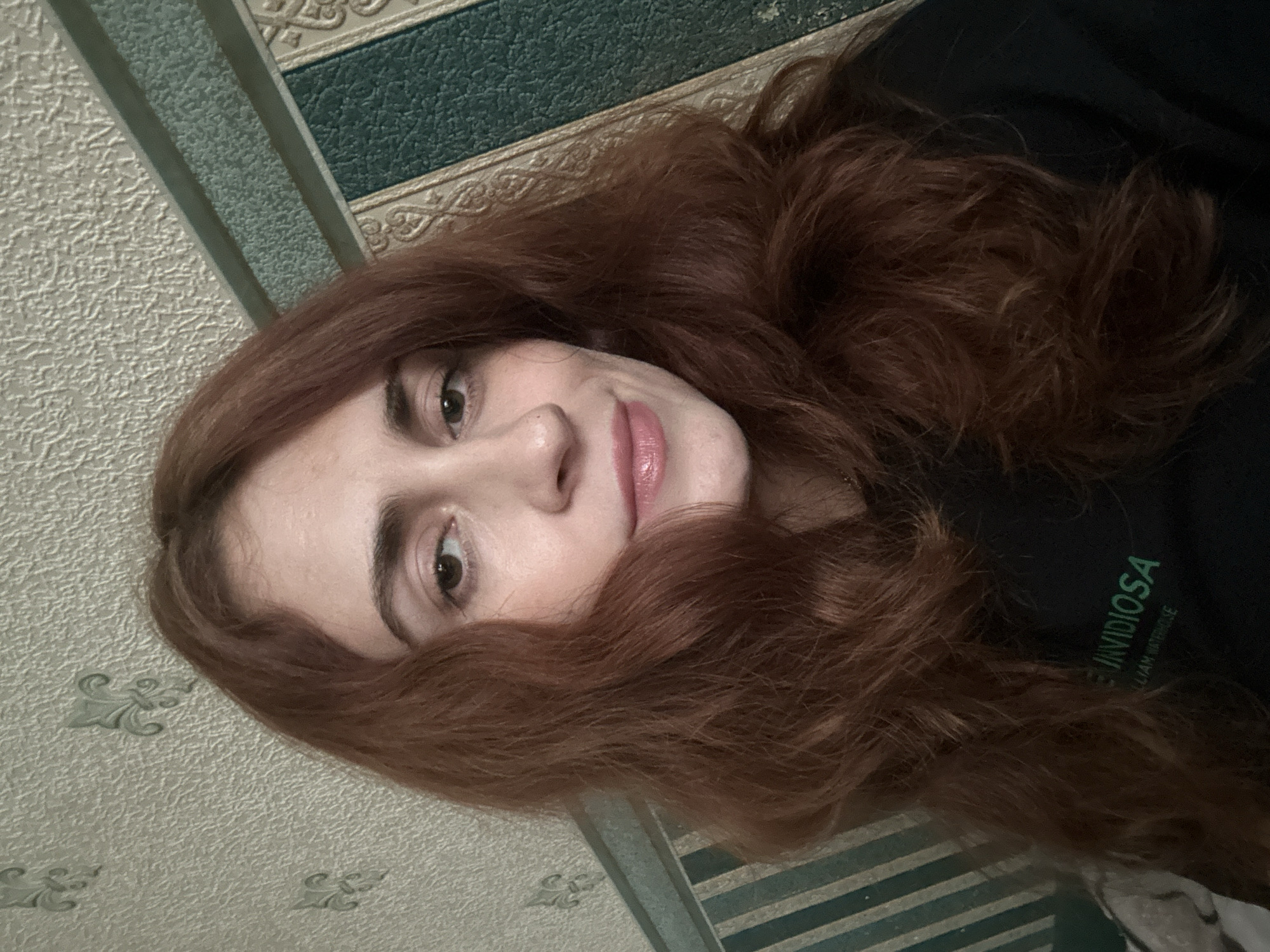 The image is a close-up selfie of a young woman with reddish-brown, wavy hair. She is looking directly at the camera with a slight smile. Her skin is fair, and she is wearing subtle makeup, including lipstick. She is wearing a dark-colored t-shirt with green text visible on the chest, which appears to read "UNVIDIOSA" and "LLIAM WATERHOUSE." The background consists of textured wallpaper with a repeating pattern of what looks like fleur-de-lis motifs, and a section with green and gold striped accents. There is no indication of the city of Togliatti, Russia, in the image.