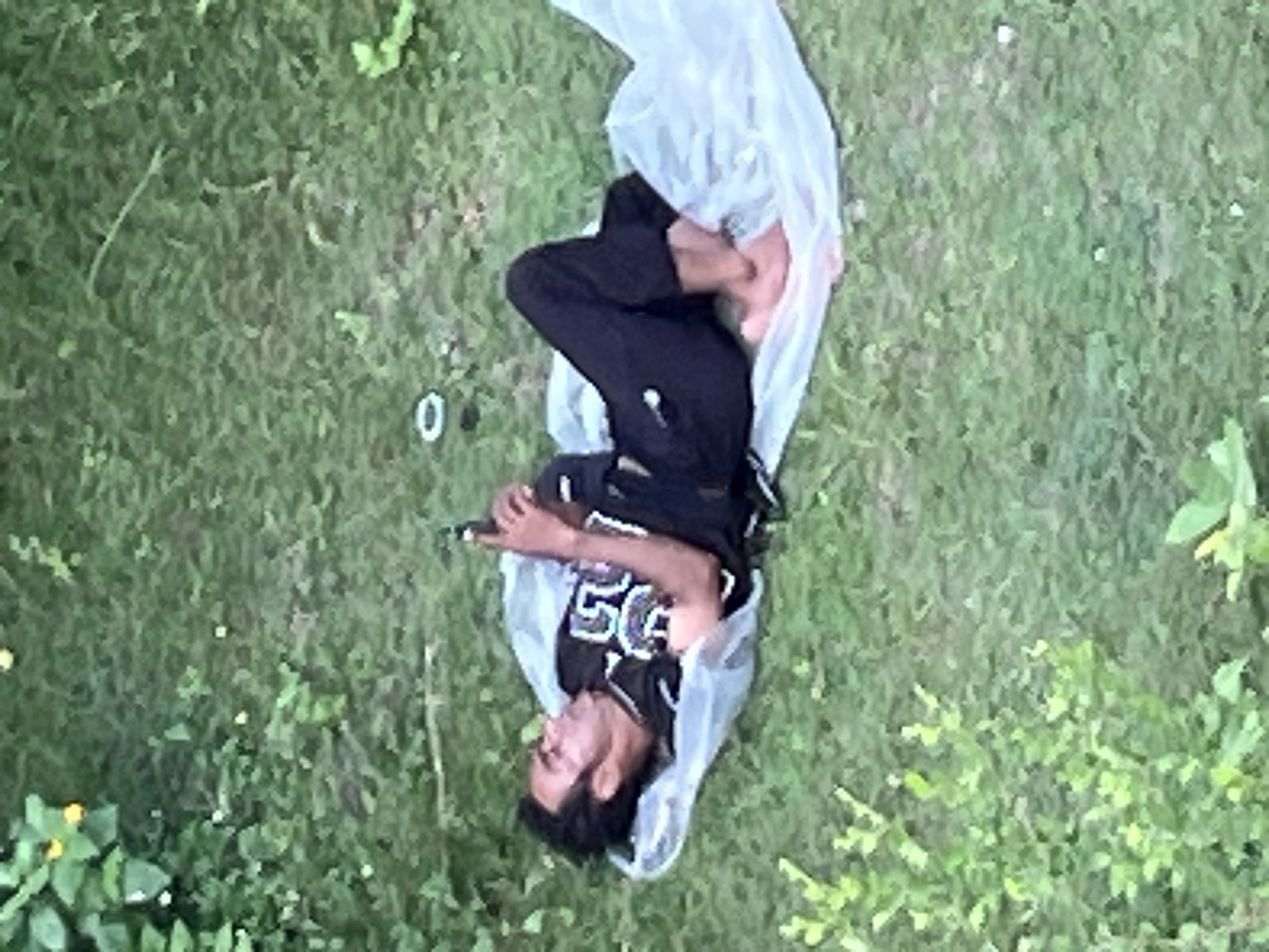 A man is lying on his back on a grassy surface. He is wearing a black shirt with the number "50" on it, black pants, and has bare feet. A semi-transparent white plastic material partially covers him. He appears to be holding a small object. The background is primarily green vegetation. A small white ring-shaped object is visible near the man. The location is in the city of Unknown, Unknown.