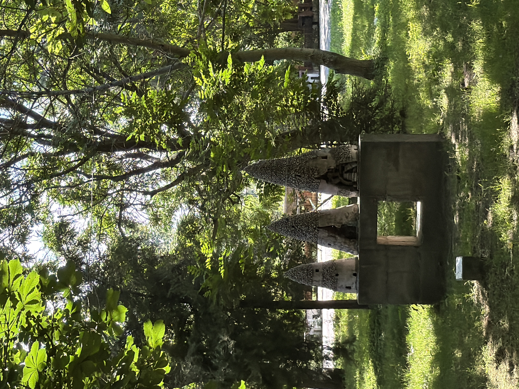 This image captures a sunlit park scene in Pazardzhik, Bulgaria. In the foreground, a low, concrete rectangular structure with a central opening sits on a patch of grass. To the right of this structure are three decorative cone-shaped objects, each adorned with what appear to be small stones and featuring openings that suggest miniature dwellings. These objects are positioned on a slightly raised concrete platform. The background is dominated by lush green trees, some with dense foliage and others with more exposed branches, indicating a park or garden setting. Sunlight filters through the leaves, creating dappled light on the grass. In the distance, beyond the trees, there are hints of structures and pathways, suggesting the presence of a larger park or possibly the edge of a town. There are no people visible in the image.